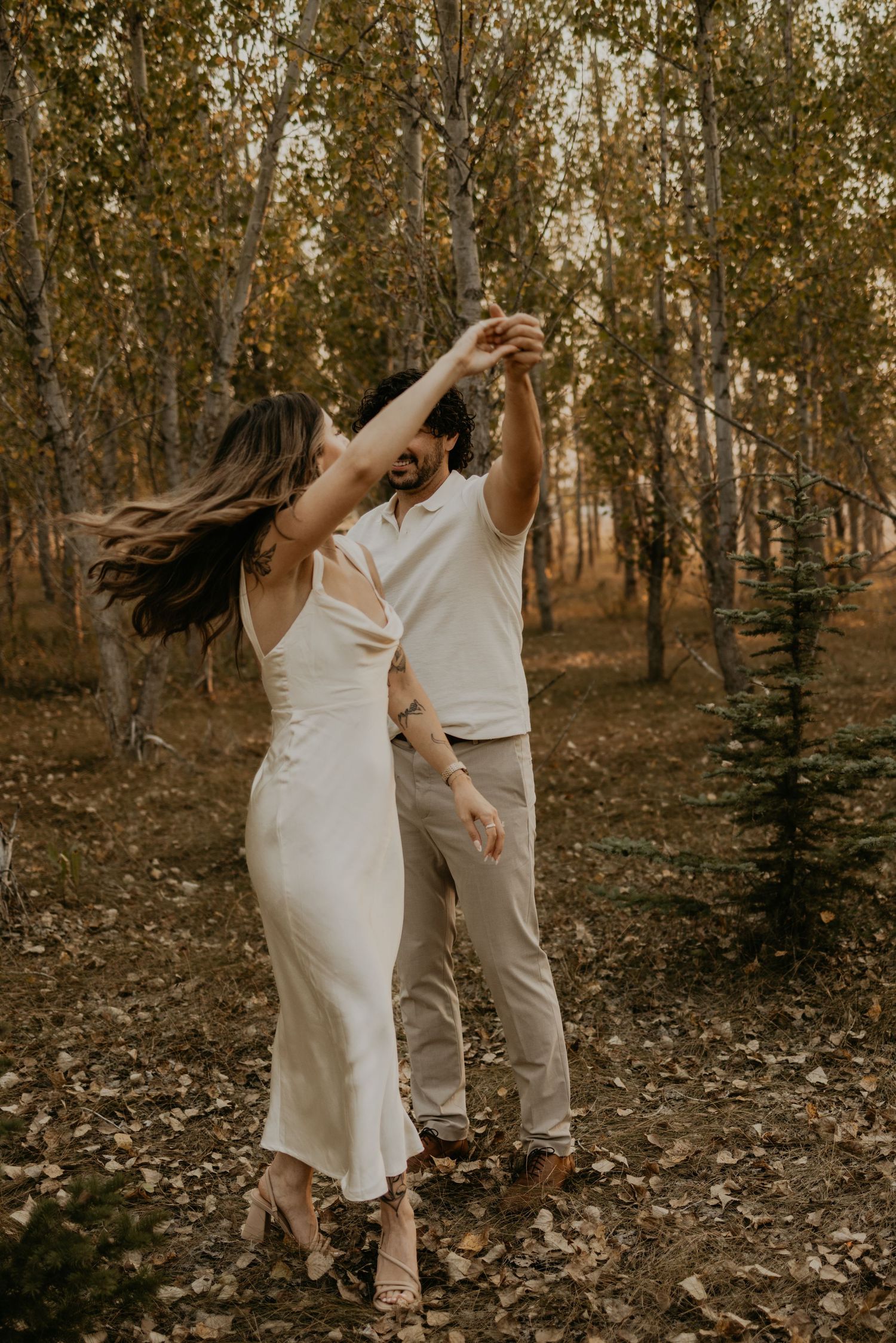 A couple in white and beige attire dances playfully in a wooded area during golden hour.