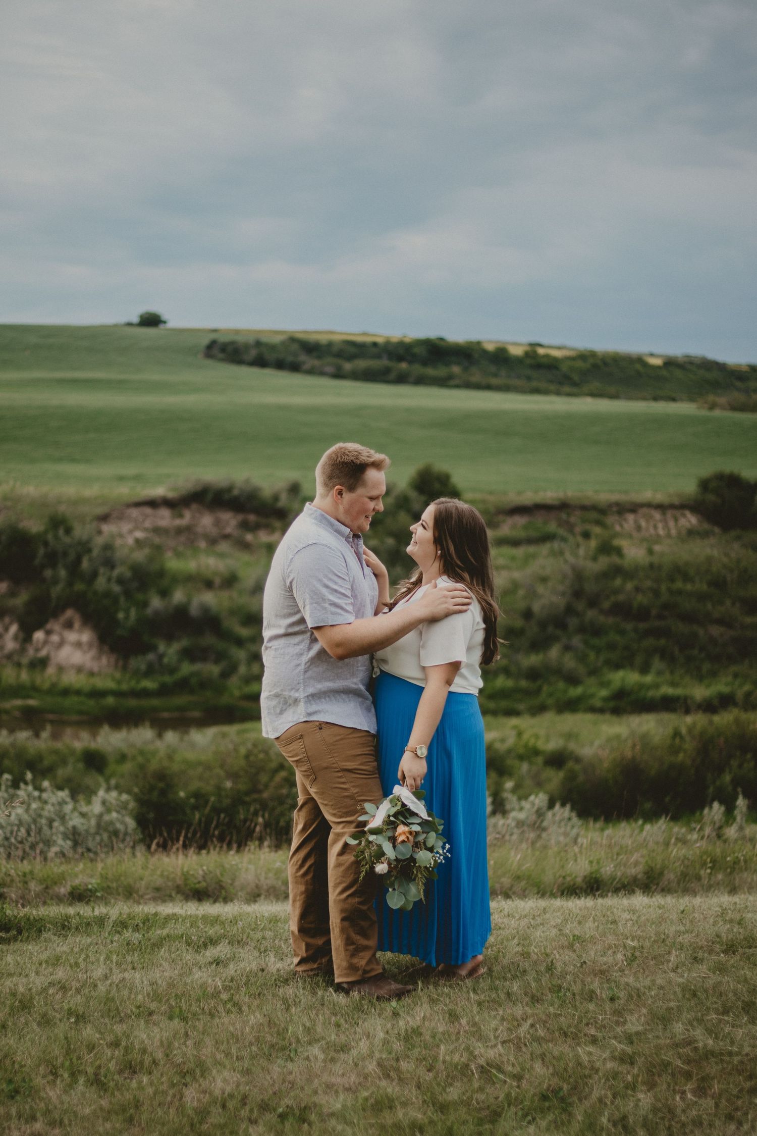 Couple stands close together in meadow with blue skirt contrasting against green landscape.