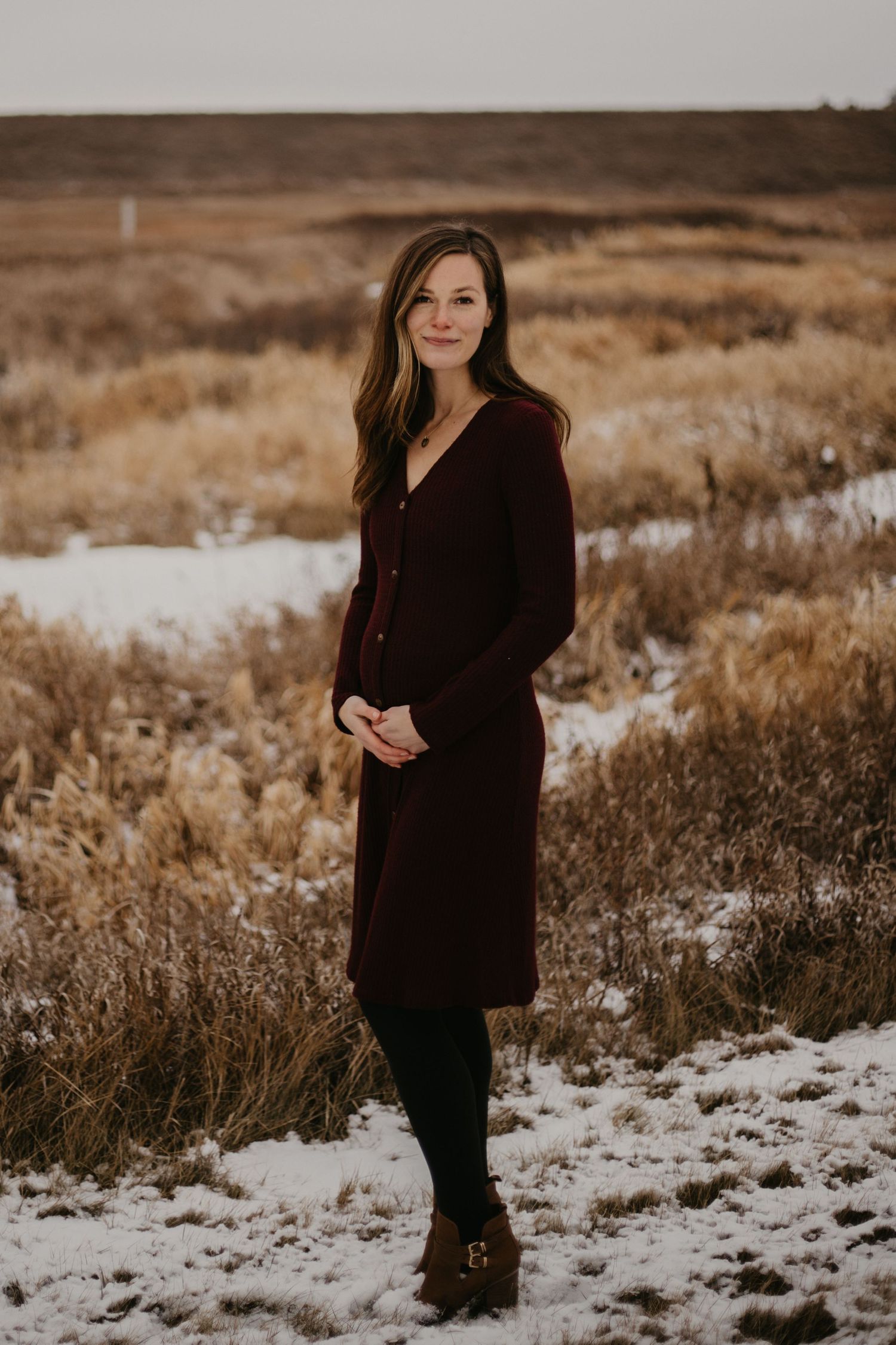 A person in a long burgundy cardigan poses in a snowy winter field.