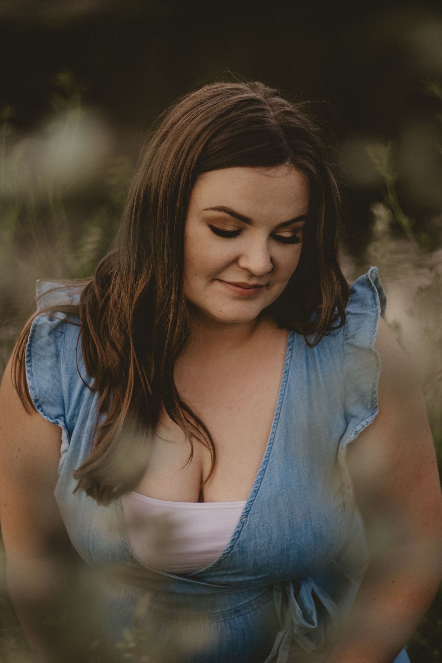 A woman in a blue denim dress smiles while looking down in a candid outdoor portrait.