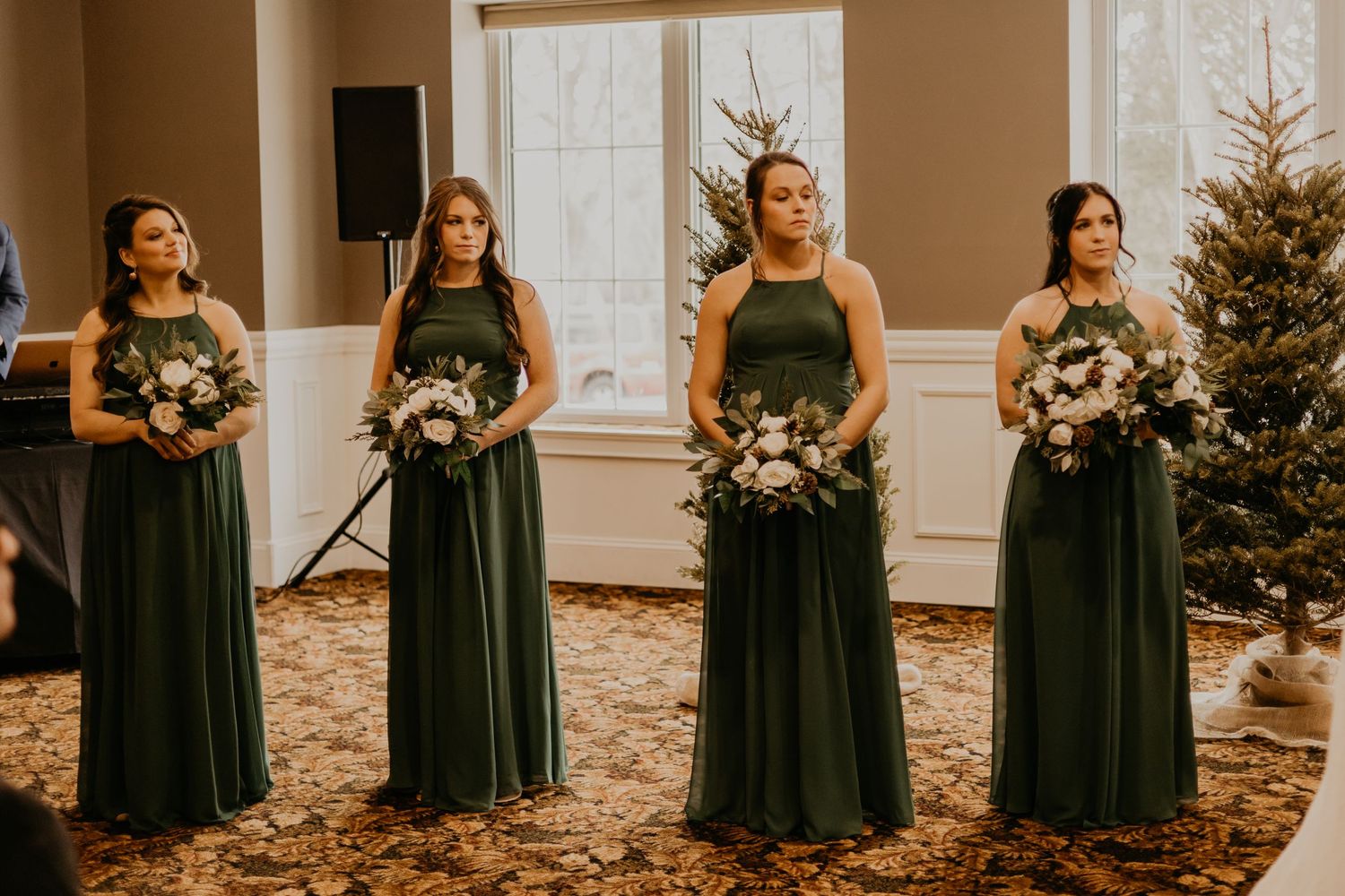 Bridesmaids in emerald green floor-length gowns hold white bouquets during indoor wedding ceremony.