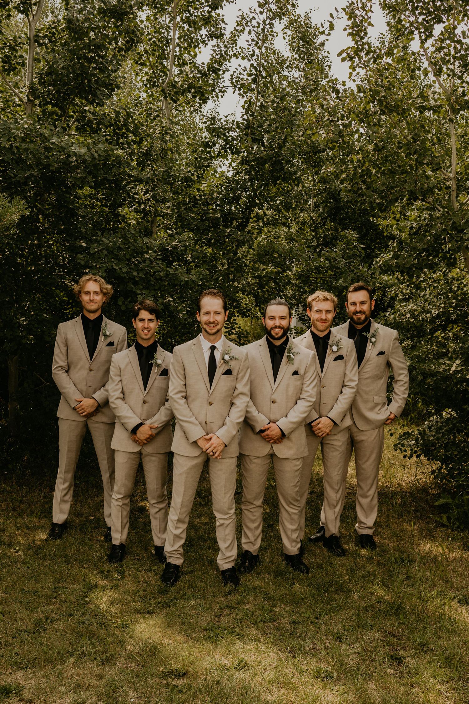 Groomsmen in beige suits stand together for formal photo against leafy green background.