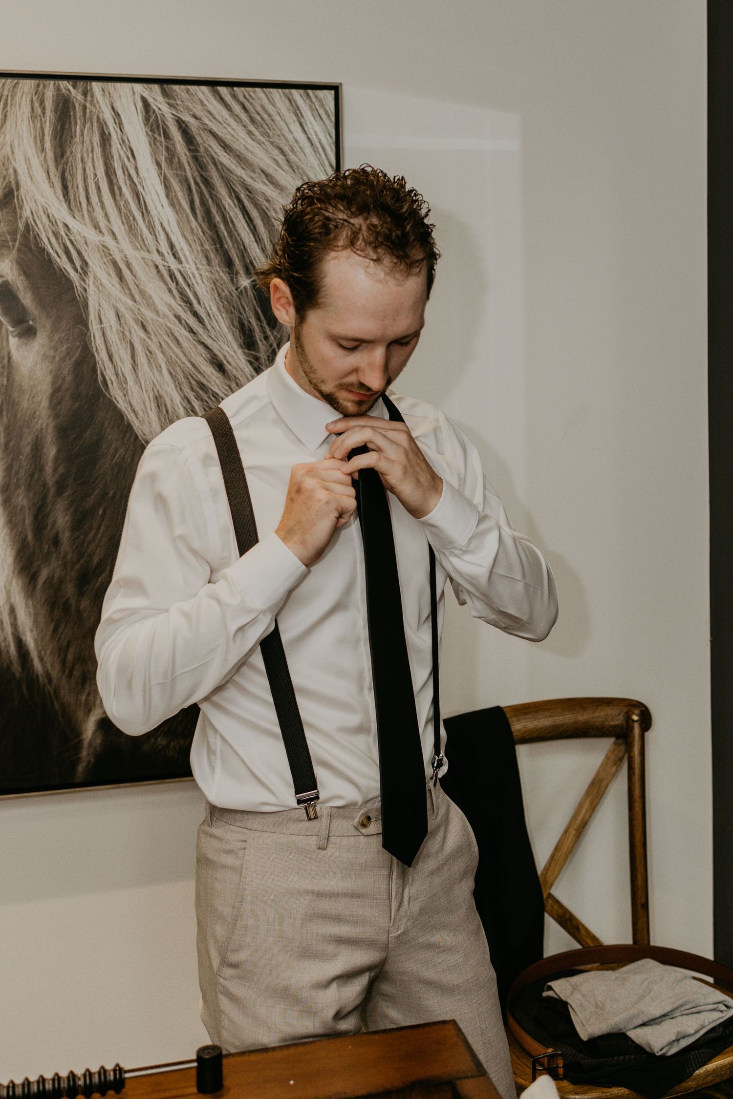 A person adjusts their tie while wearing suspenders and a white dress shirt.