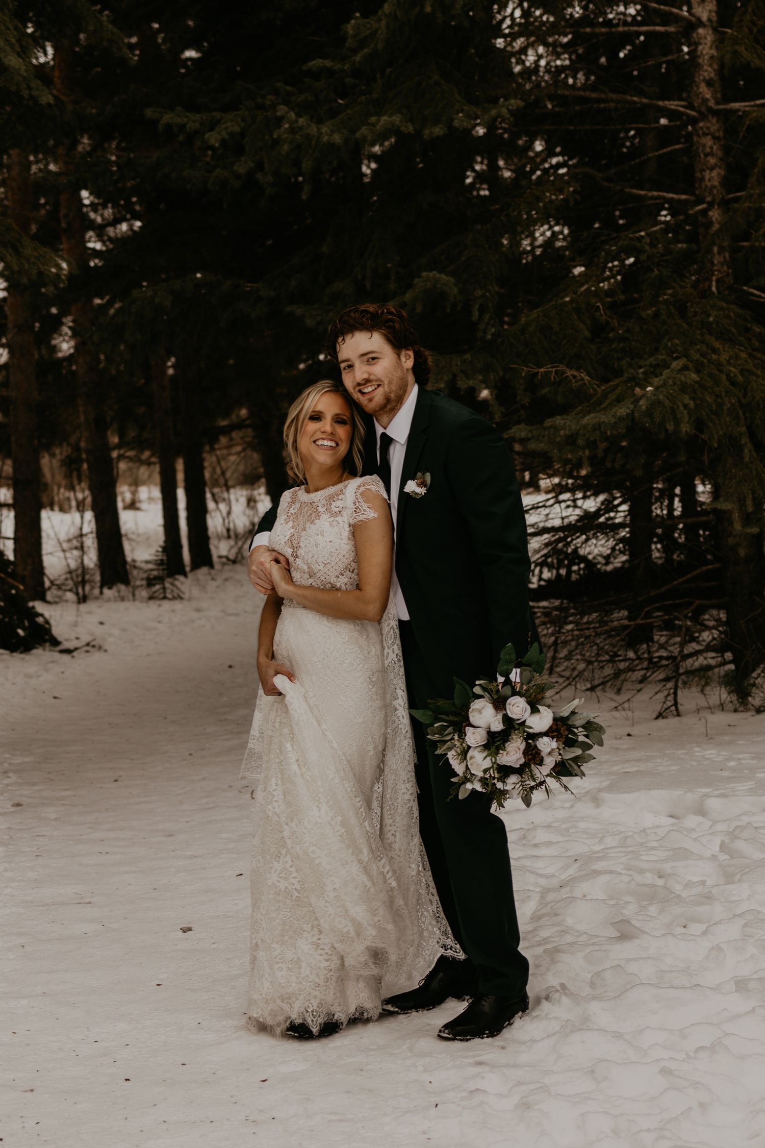 A newlywed couple poses in a winter forest landscape with snow-covered ground and evergreen trees.