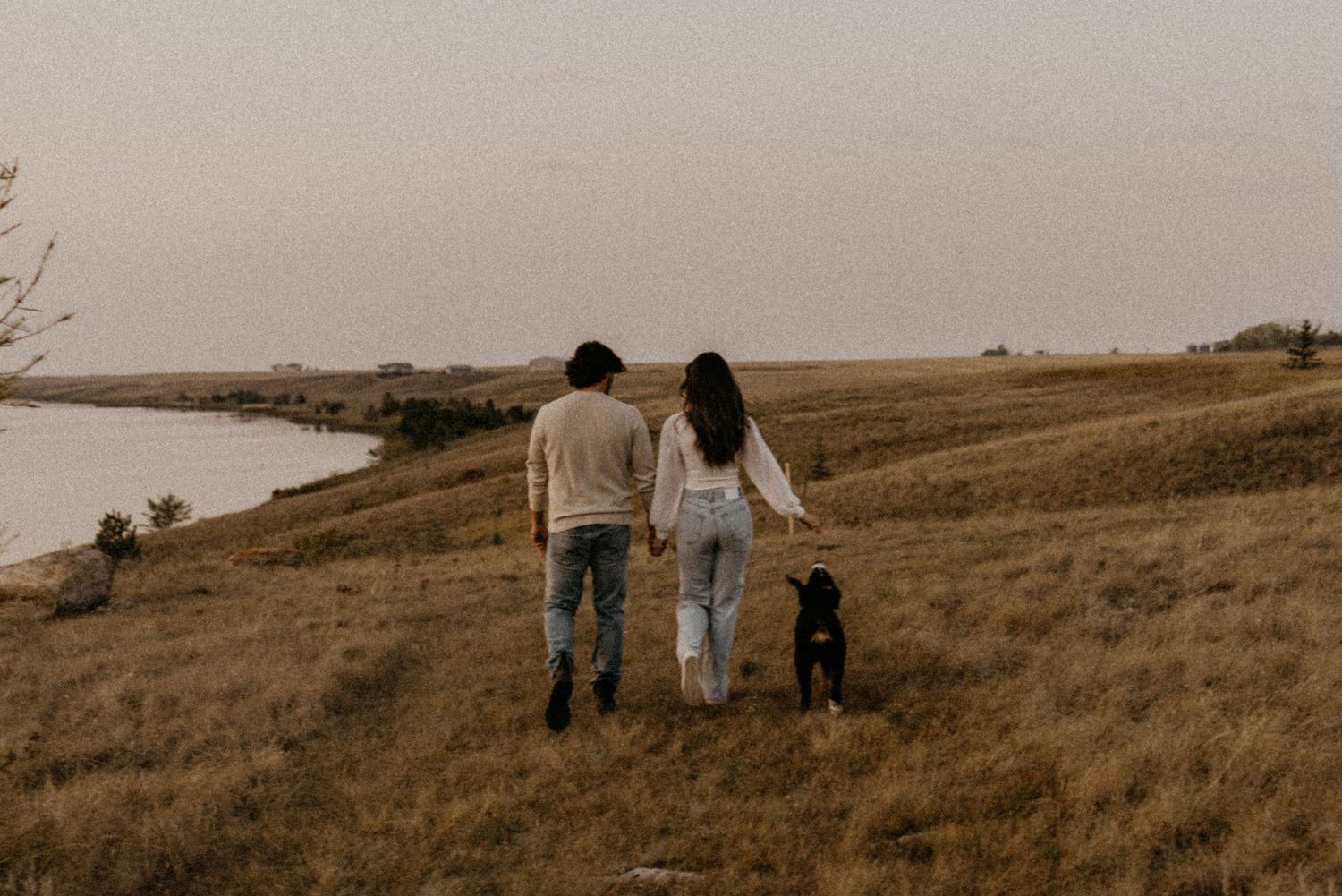 A couple and their black dog walk along a hillside overlooking a serene lake at sunset.