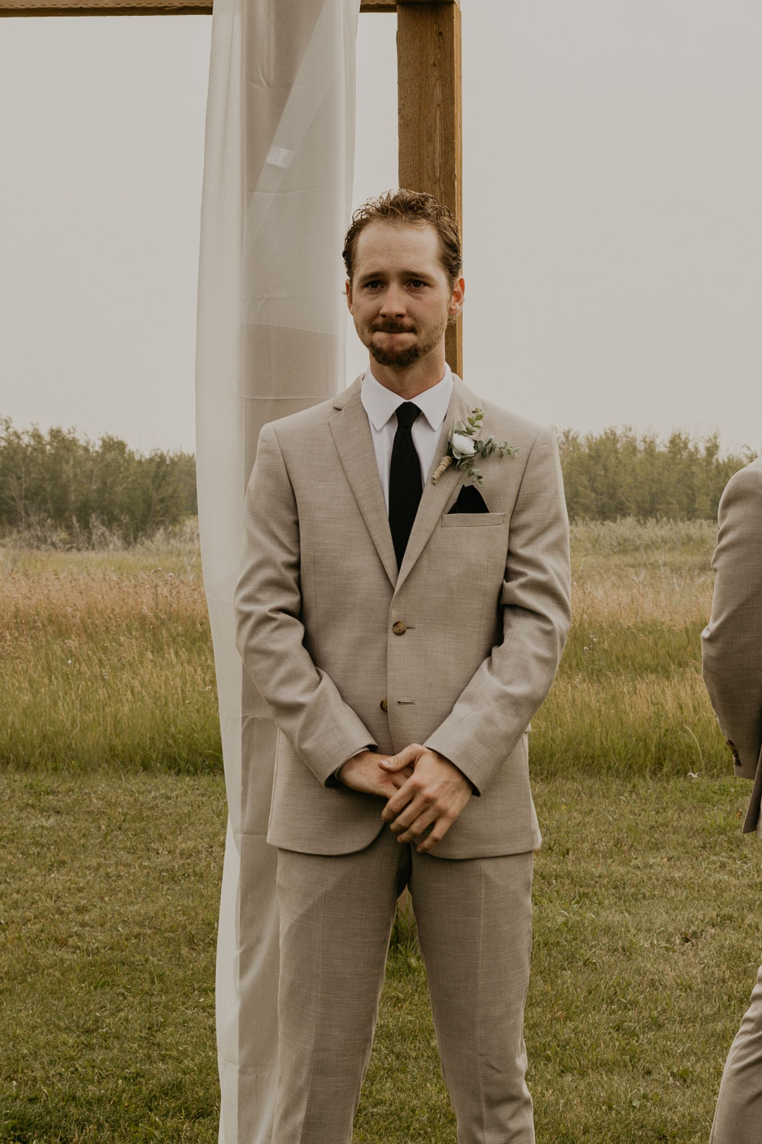A groom in a beige suit and black tie stands at attention during an outdoor wedding ceremony.