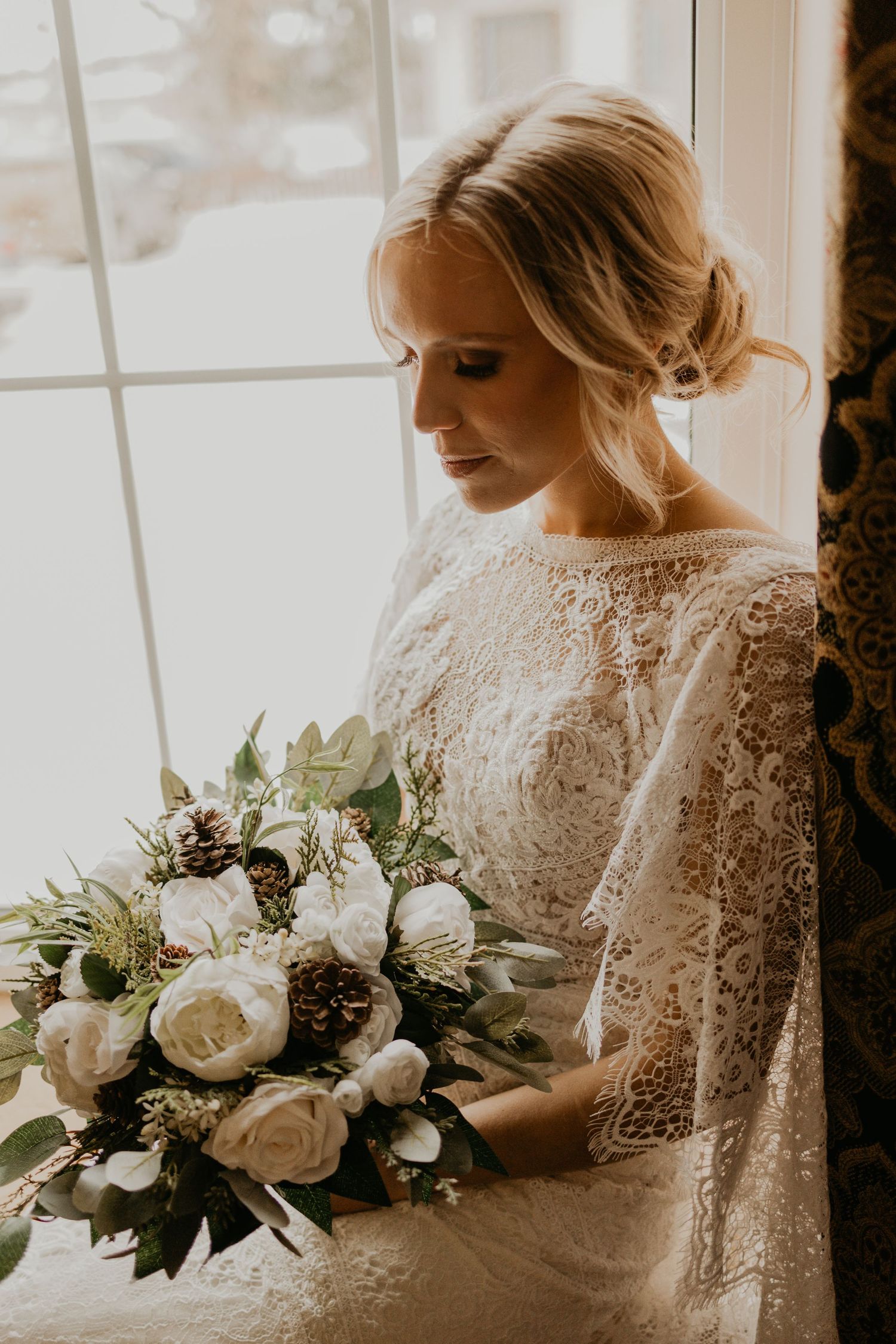 A wedding bouquet with white roses and pinecones is held against a lace dress by a window.