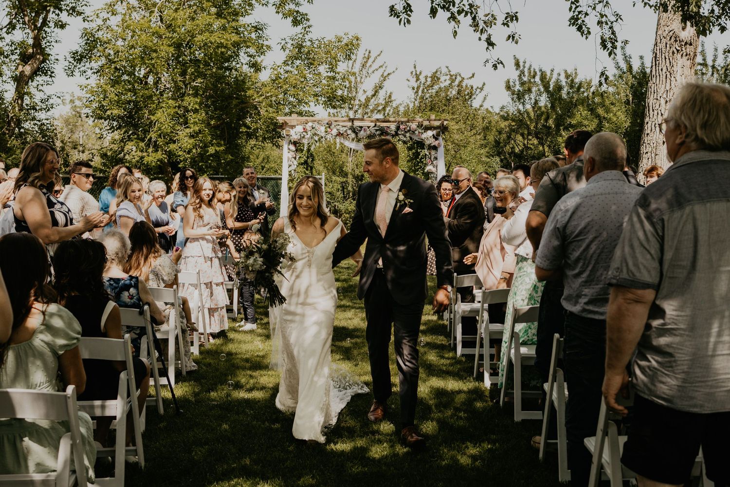 Wedding couple walking down the aisle while guests cheer during an outdoor garden ceremony.