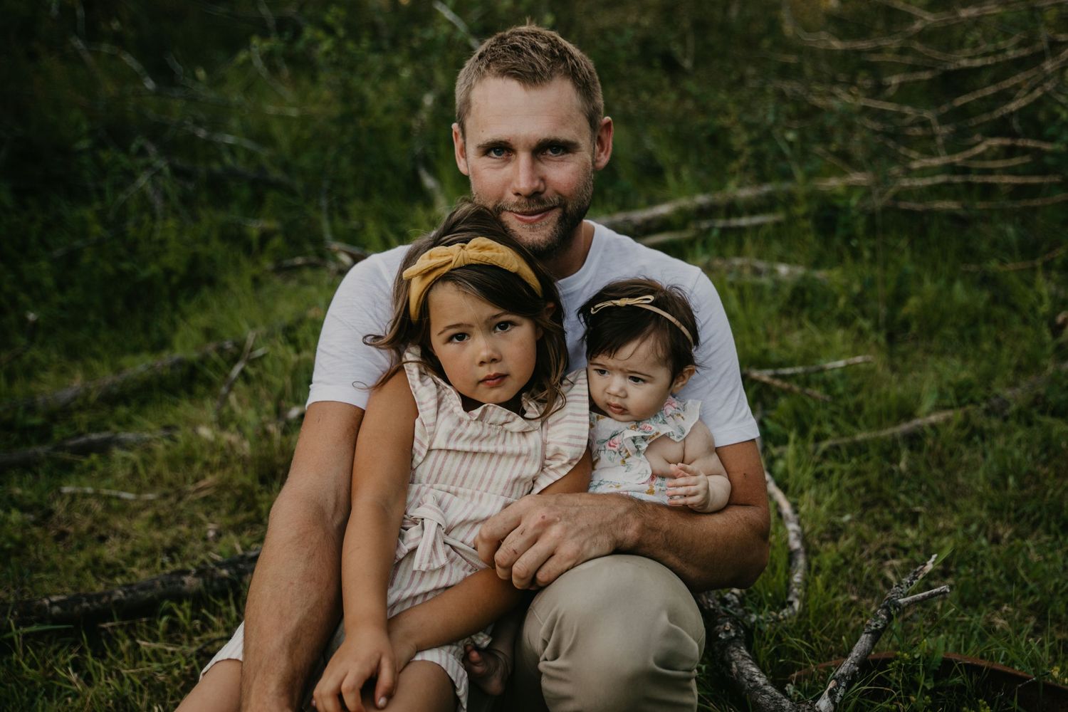 A parent and two small children sit together outdoors in a natural setting during golden hour.