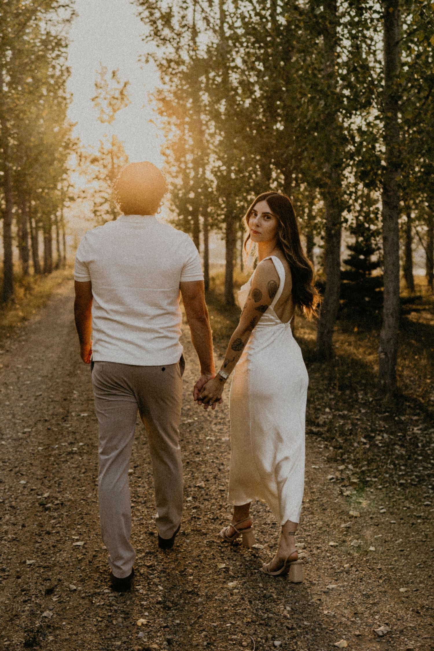 Couple walking hand in hand down a tree-lined path at sunset in coordinated white and beige outfits.