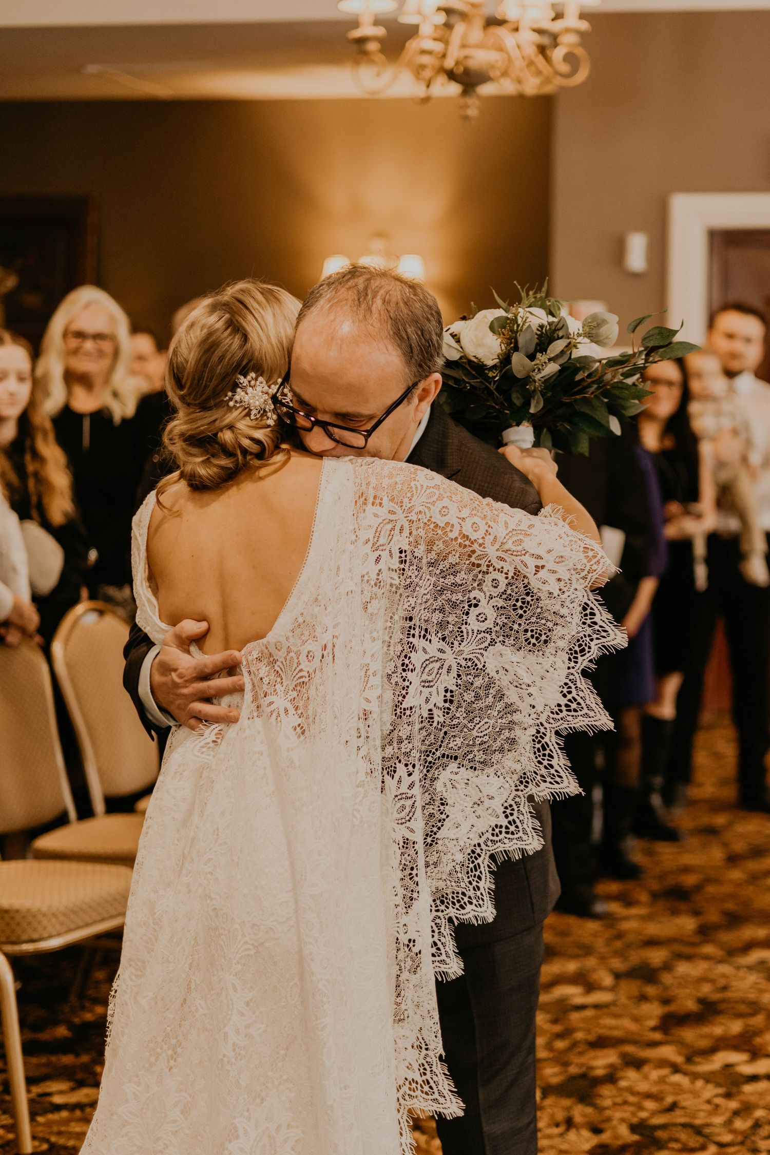 An emotional embrace during a wedding celebration in a room with chandelier lighting and gathered guests.