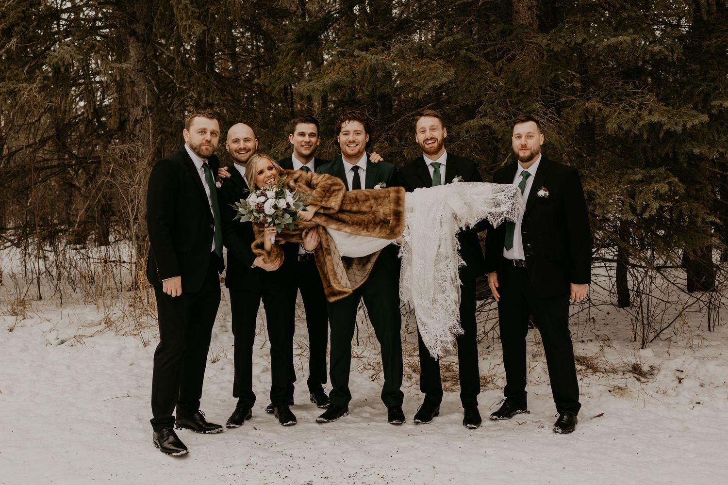 Groomsmen in black suits pose with decorative winter props against snowy forest background.