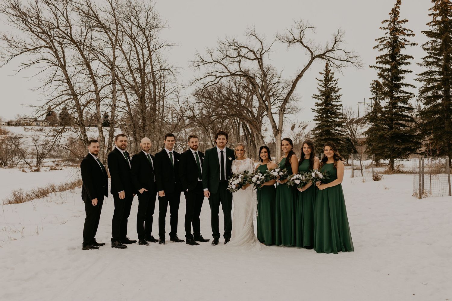 Wedding party in black suits and emerald dresses poses together in a snowy outdoor setting.