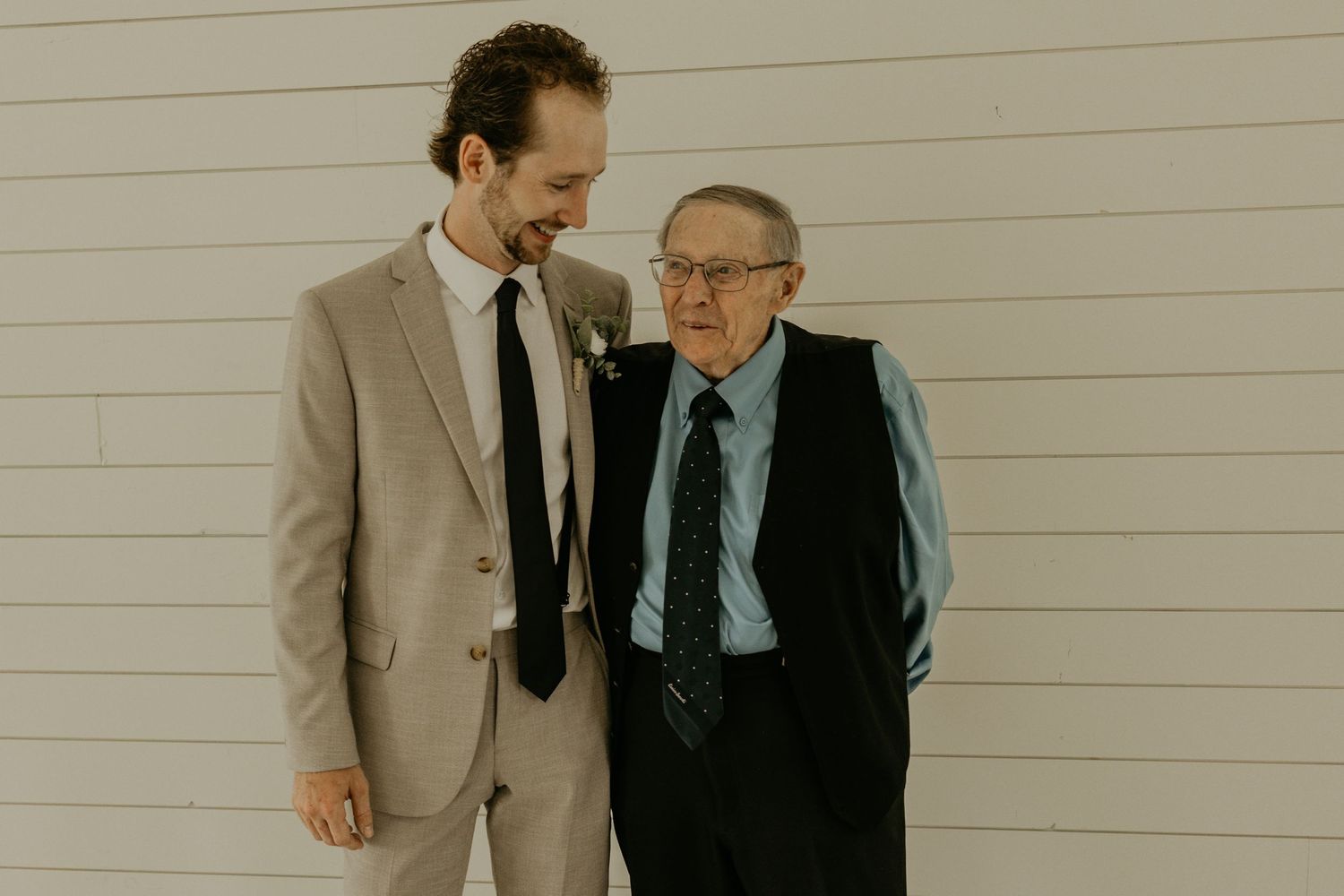 Two generations pose together at wedding, one in tan suit and other in classic black suit.