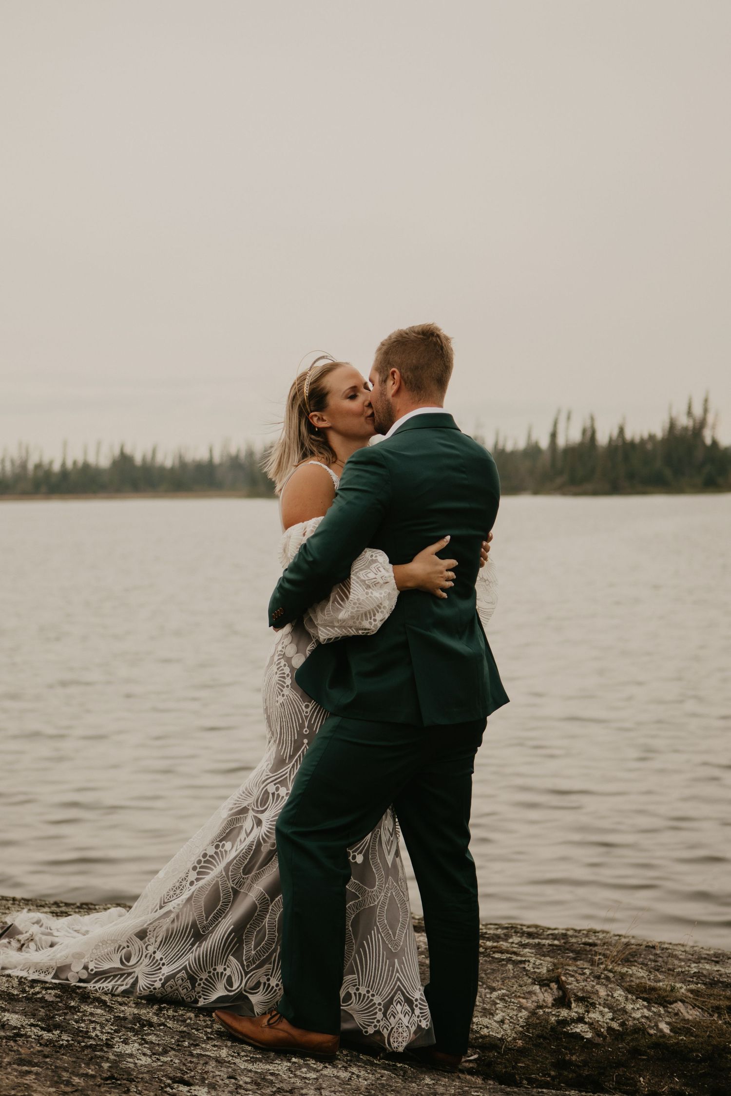 A romantic embrace between newlyweds on a rocky shoreline with a lake and forest backdrop.