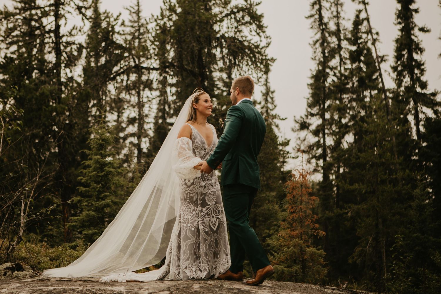 A couple shares an intimate moment on a mountain top surrounded by pine trees at sunset.