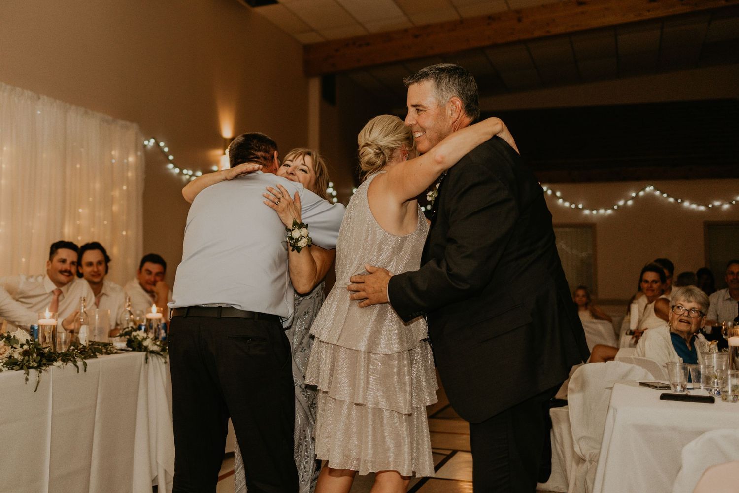 Emotional group hug during a wedding reception under string lights.