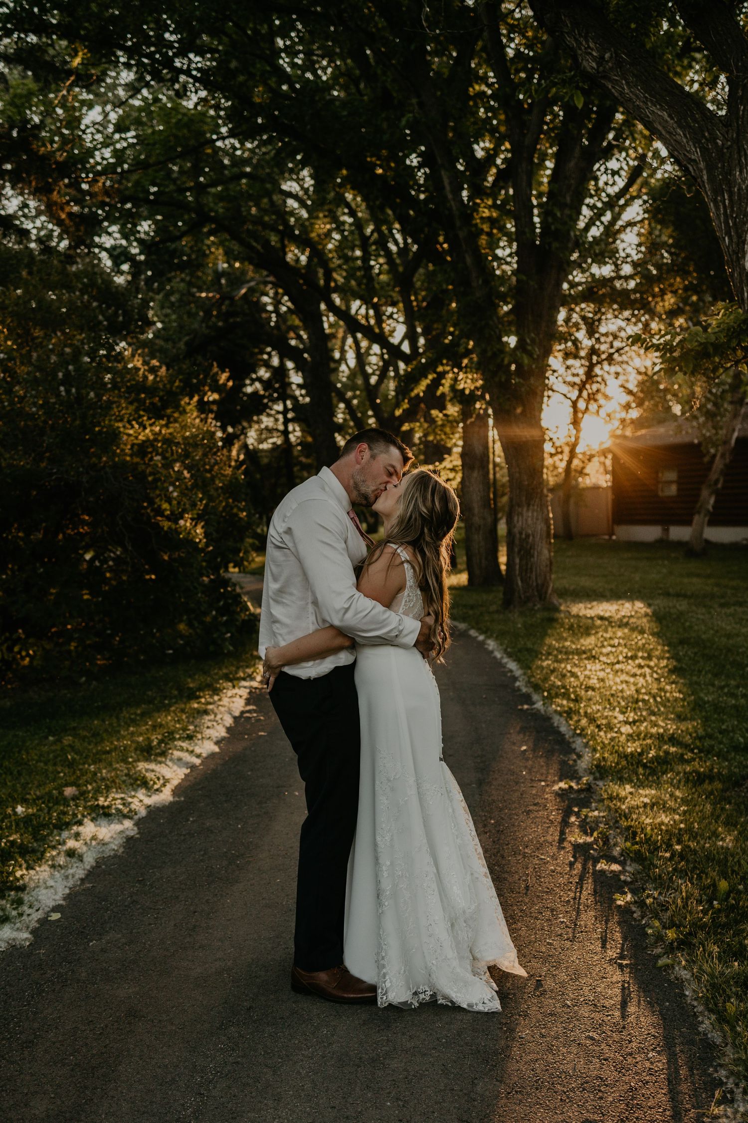 Golden hour wedding photo of couple embracing on a tree-lined pathway.