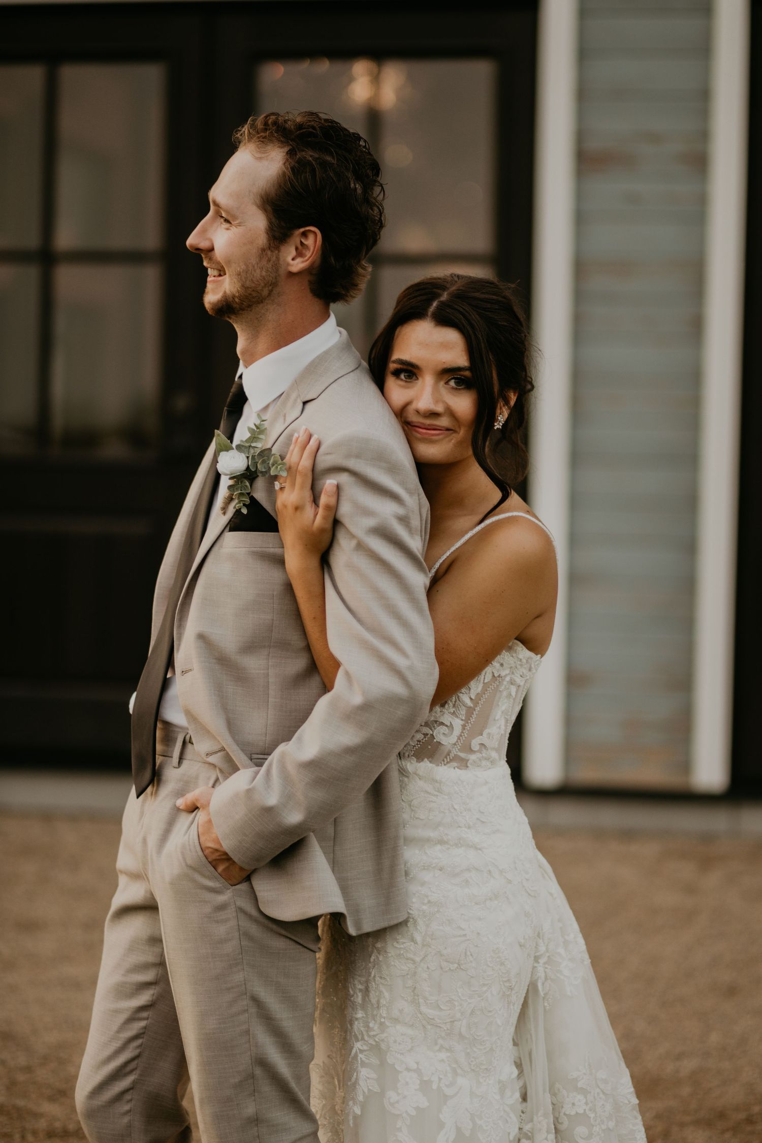 A wedding couple in tan suit and lace dress pose together outdoors.