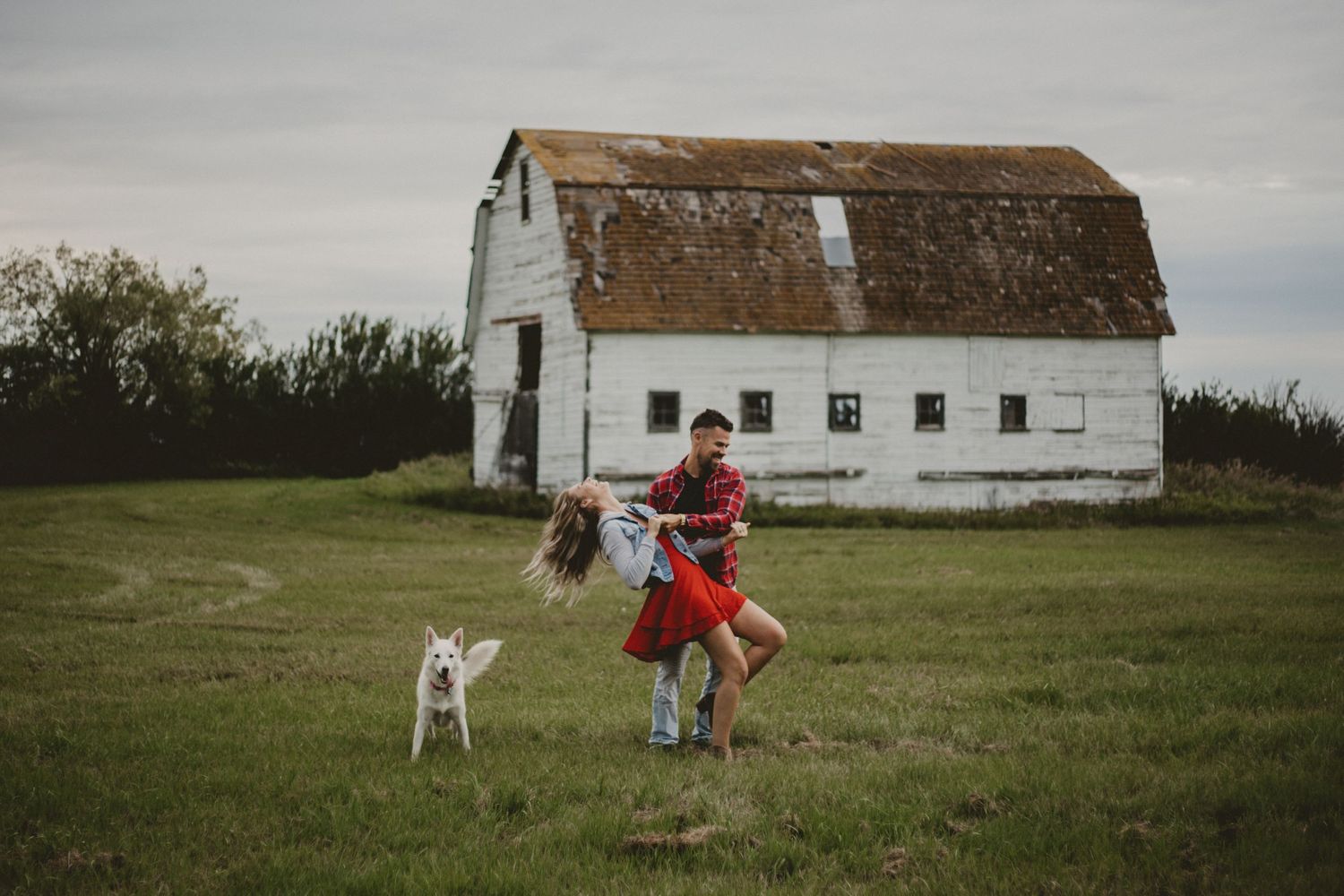 A playful scene of a couple running with their dog near an old white barn.