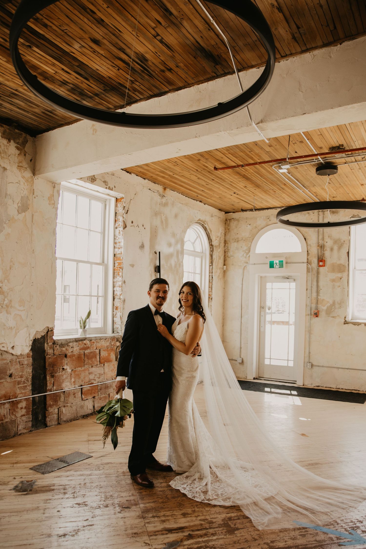 A newlywed couple embraces in a rustic venue with wooden ceiling beams and circular light fixtures.