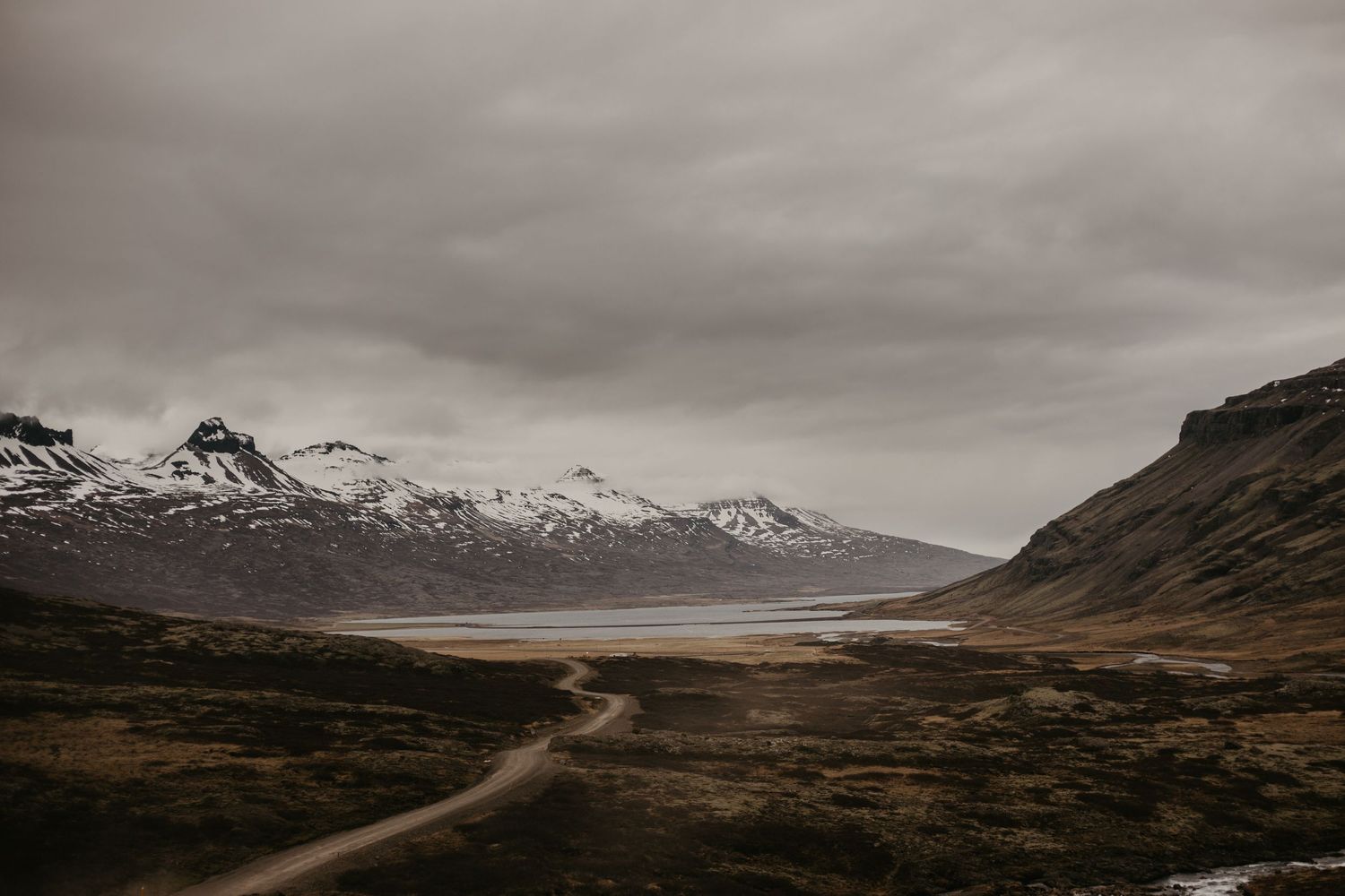 A winding dirt road leads through a dramatic mountainous landscape with snow-capped peaks.
