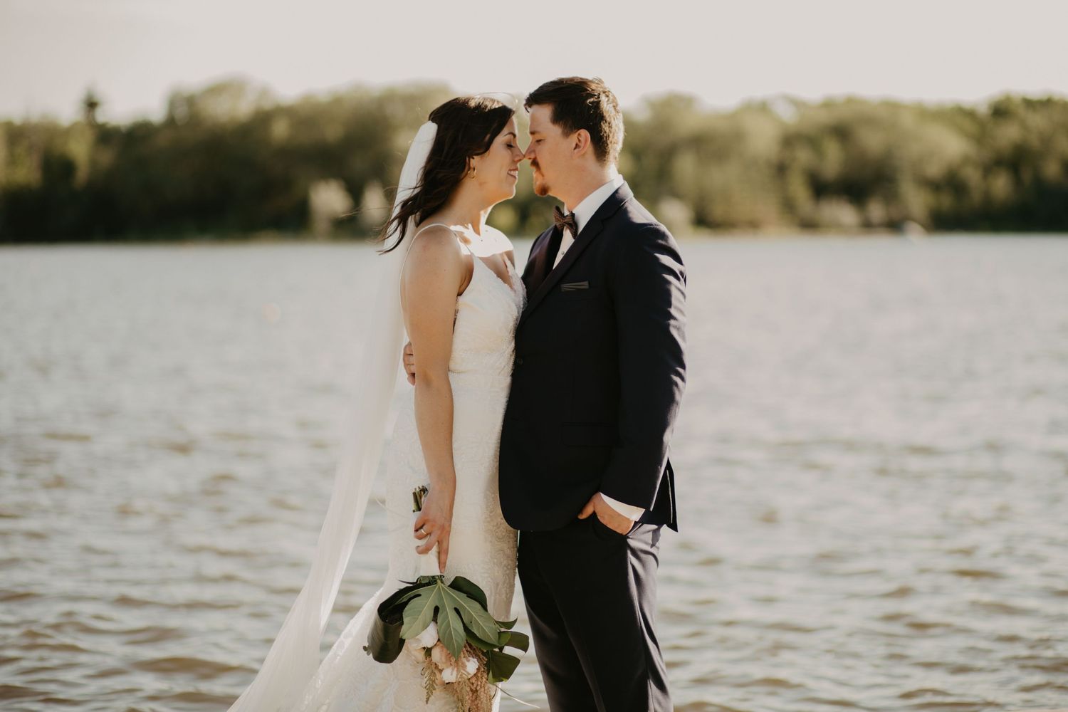 Newlyweds share intimate moment by waterfront during golden hour wedding photoshoot.