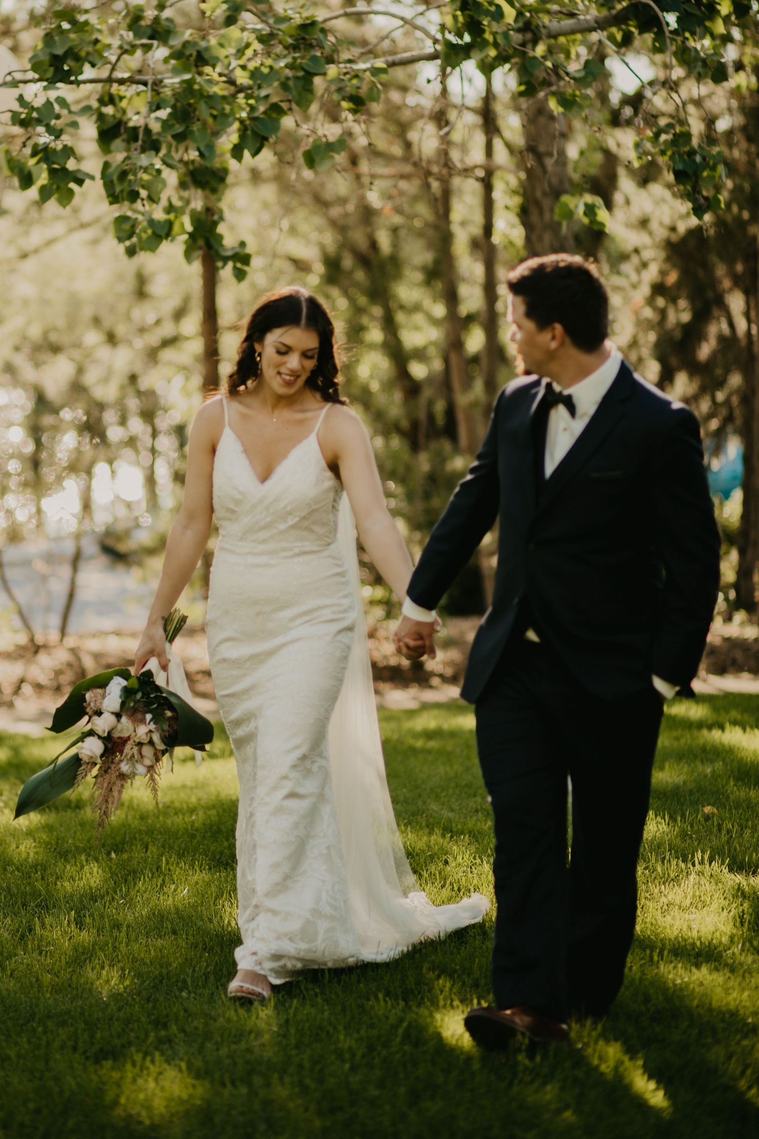 Couple walking together outdoors with the bride carrying a bouquet through a sunlit garden.