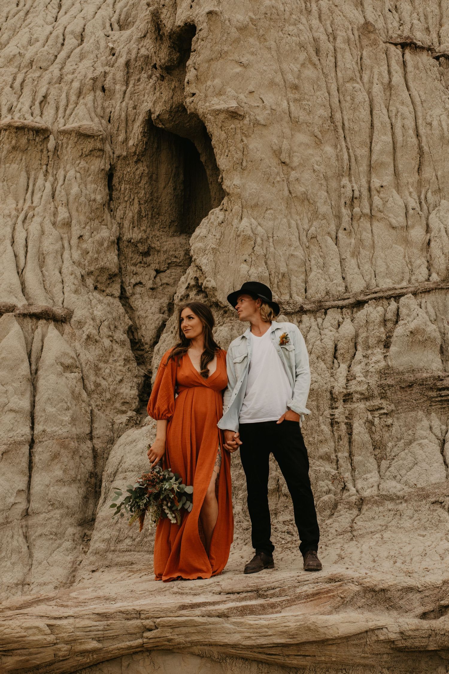 A couple stands against textured rock formations, with woman in flowing rust dress holding flowers.