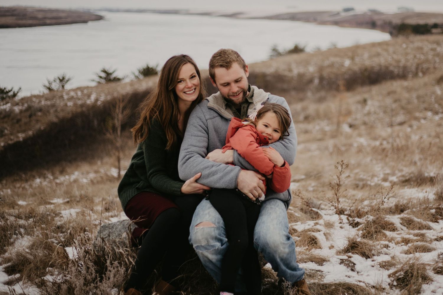 A young family sits together on rocks overlooking a serene waterfront landscape.