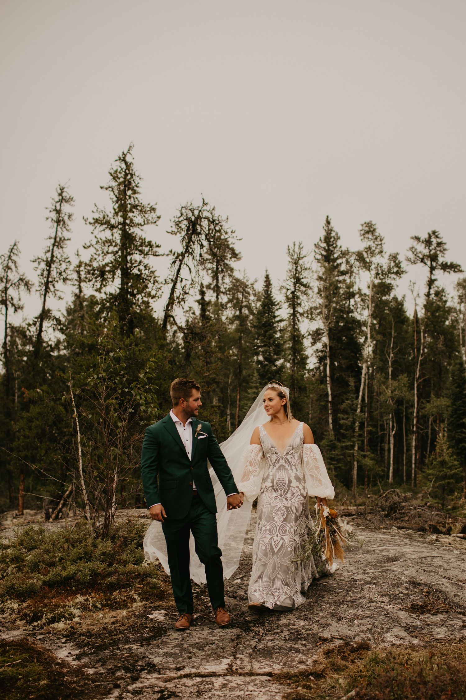 Newlyweds walk hand in hand through rugged mountain terrain surrounded by pine trees.