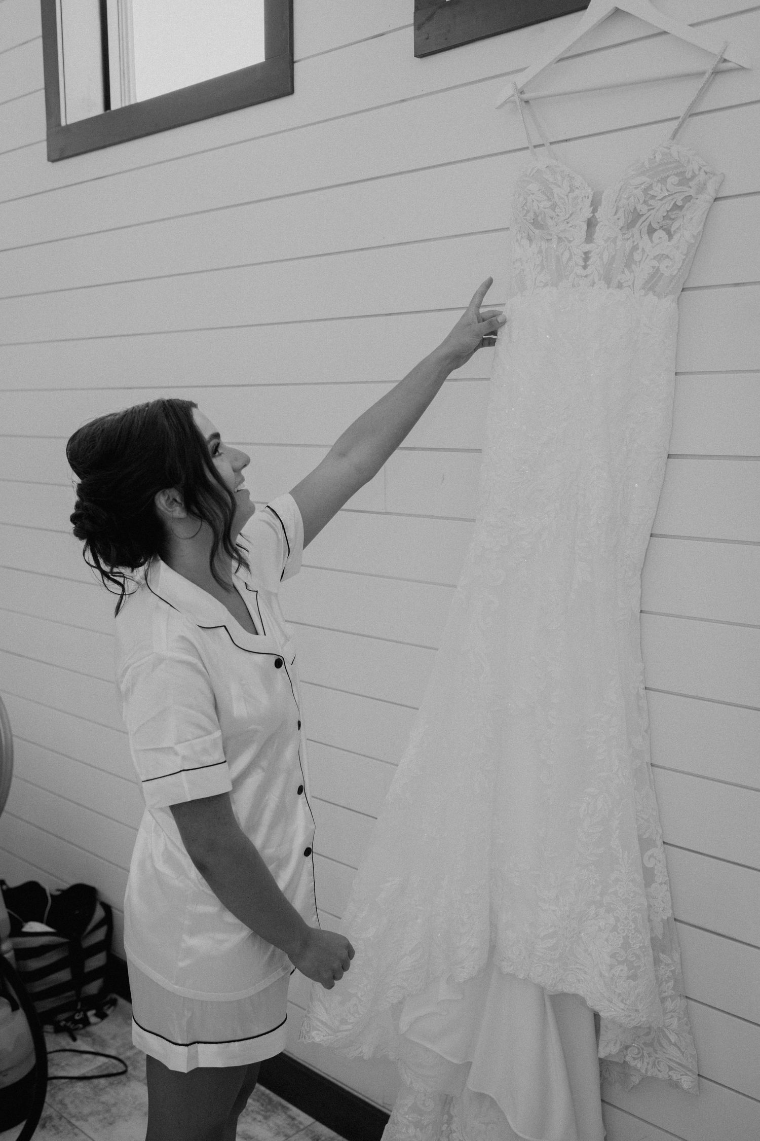 A white lace wedding dress hangs on display against a white wooden wall.