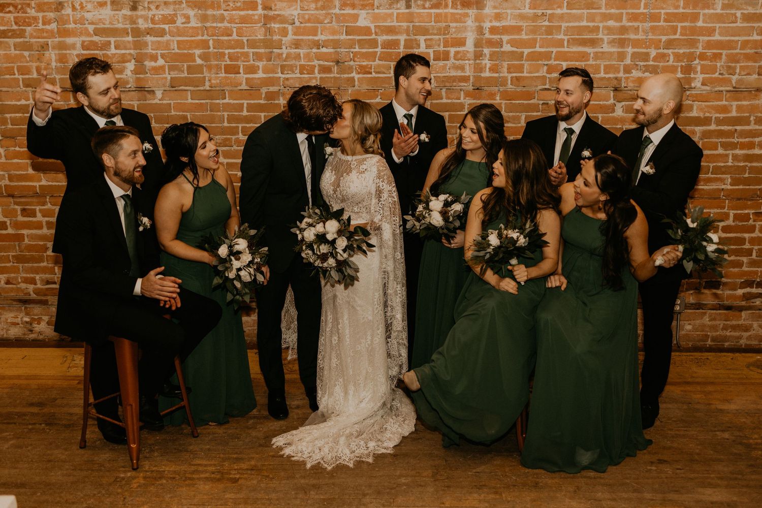 Wedding party gathered for formal photo against exposed brick wall with dark green bridesmaids dresses.