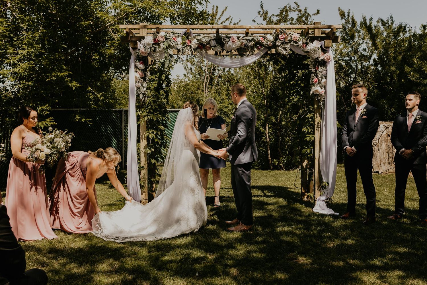 Wedding ceremony under a rustic wooden arch decorated with white fabric in a sunlit garden setting.