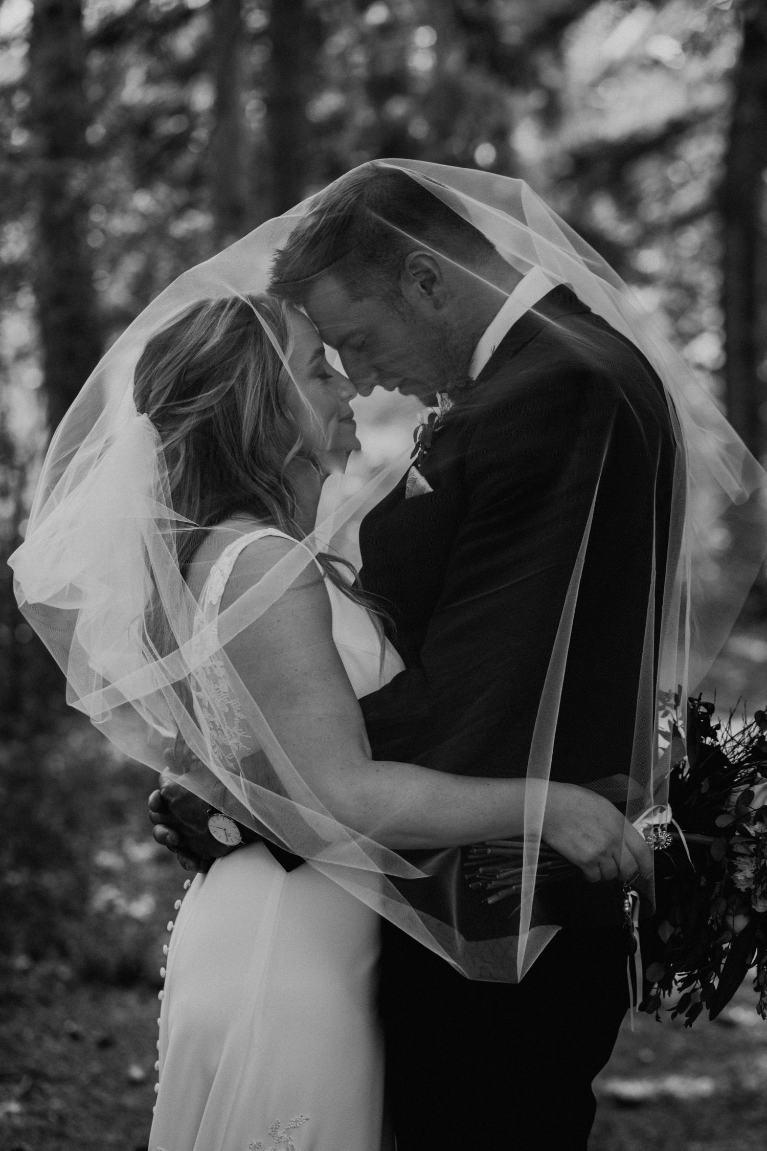 A romantic black and white portrait of a couple embracing under a flowing veil.