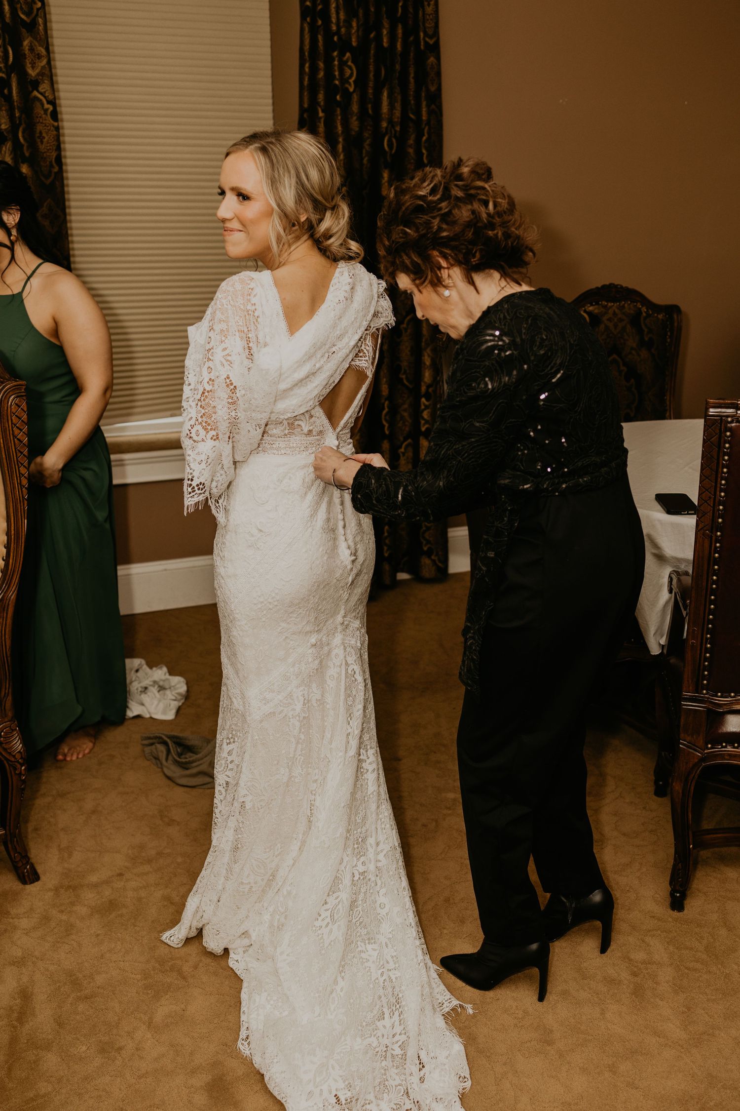 A lace wedding dress being fitted to a bride while attendants help with final adjustments.