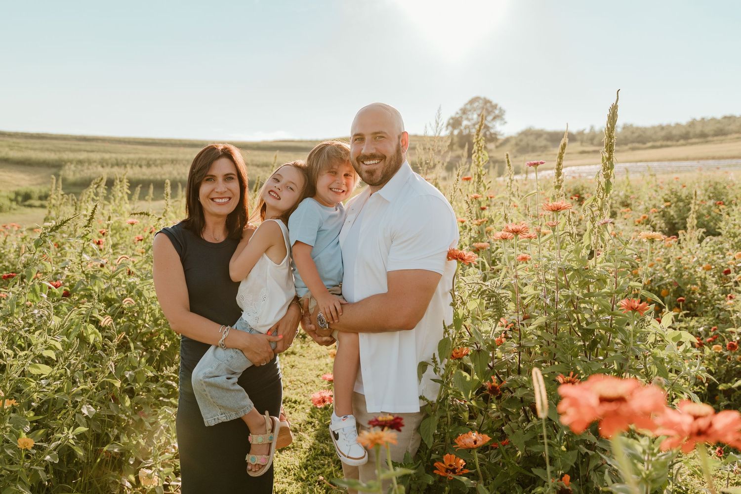 Family of four smiling in a sunny flower field with red blooms and green crops in the background.