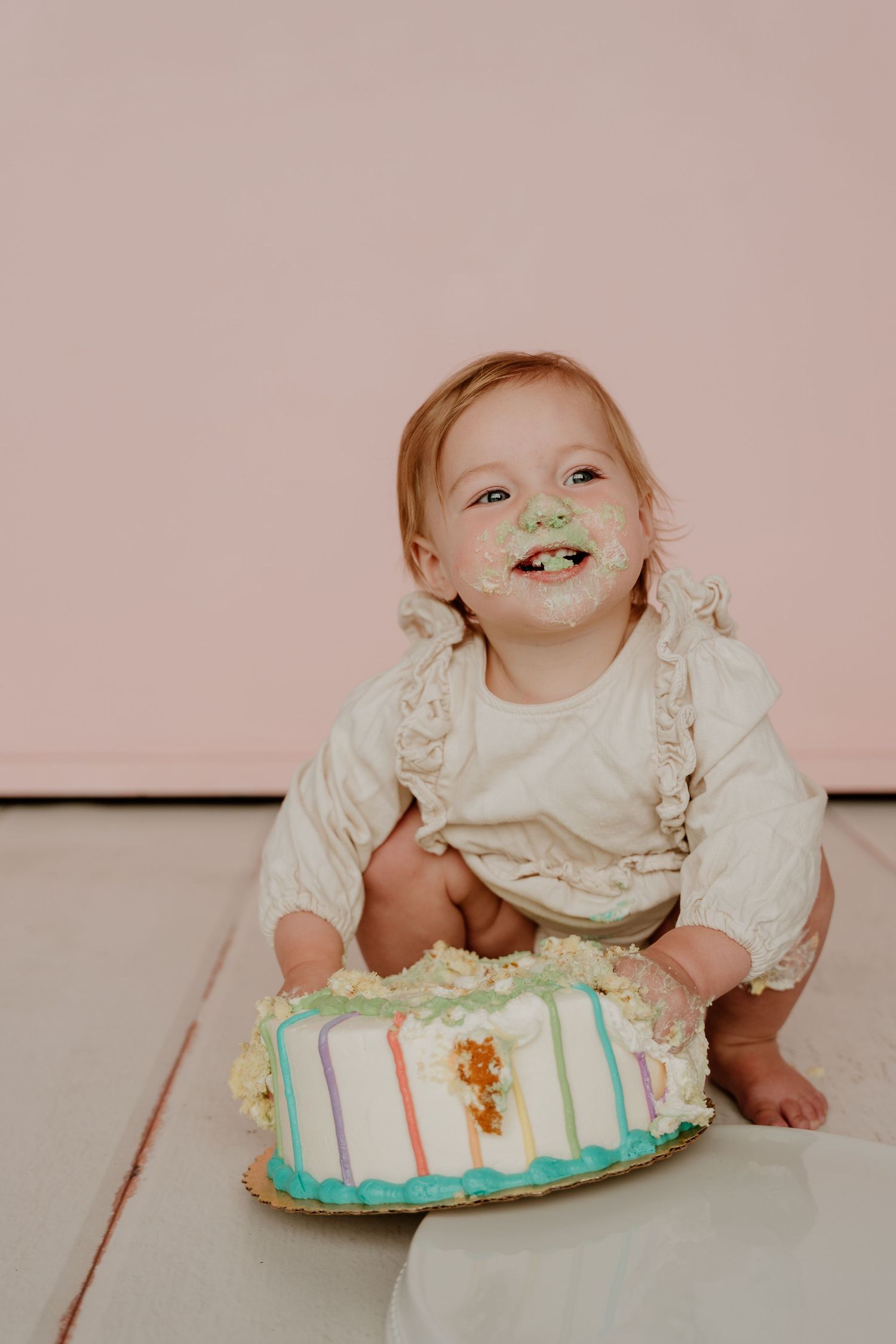 Baby in white outfit explores birthday cake during cake smash photoshoot.