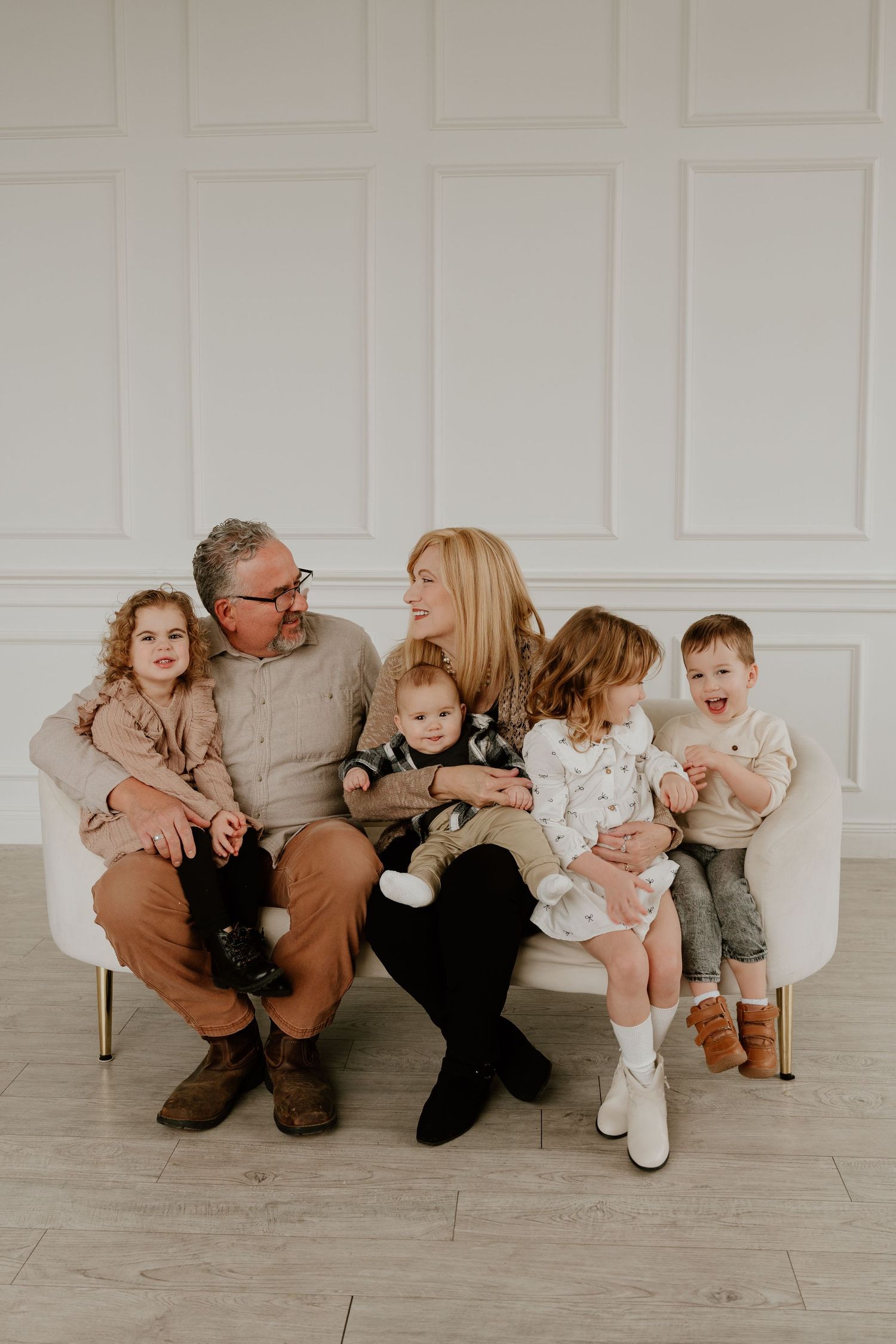 Family dressed in neutral colors sits together on white couch in minimalist studio setting with their pet cat.