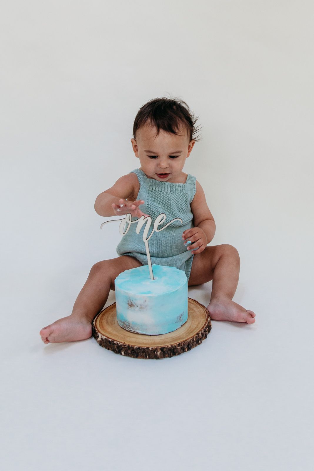 Baby in mint green outfit reaching for turquoise birthday cake on wooden slice.