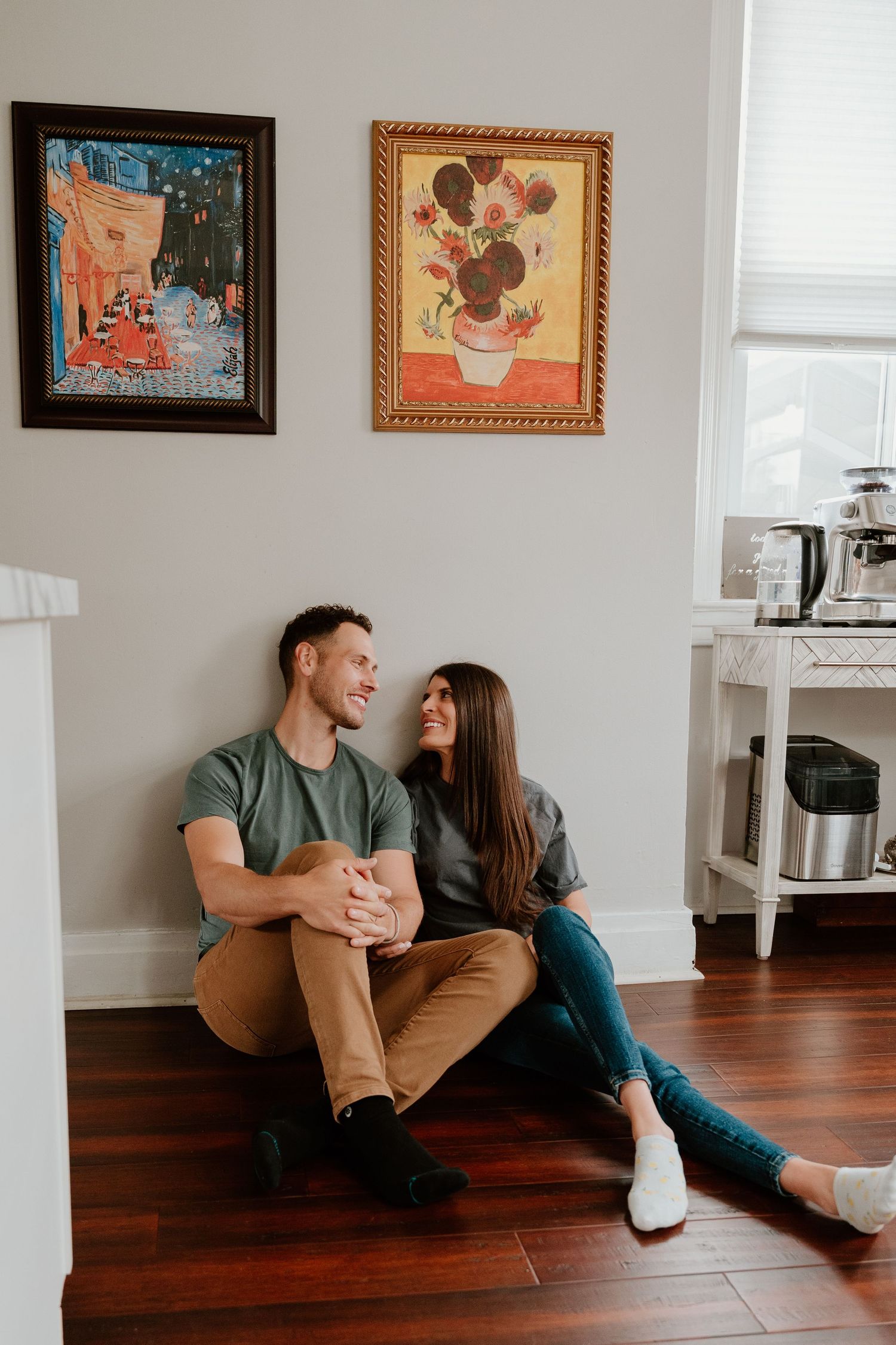 Couple sitting on hardwood floor in living room with framed artwork on wall laughing together in sequence of photos.