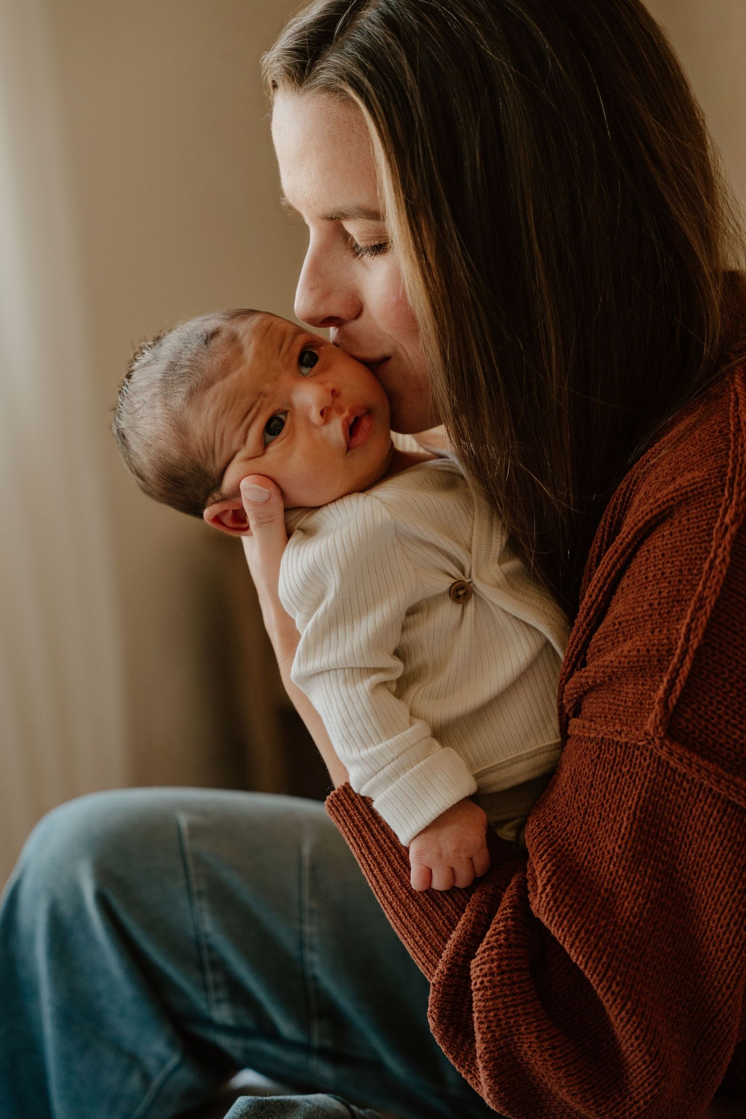 A tender moment between parent and newborn baby in warm lighting, wearing a brown cardigan and white onesie.