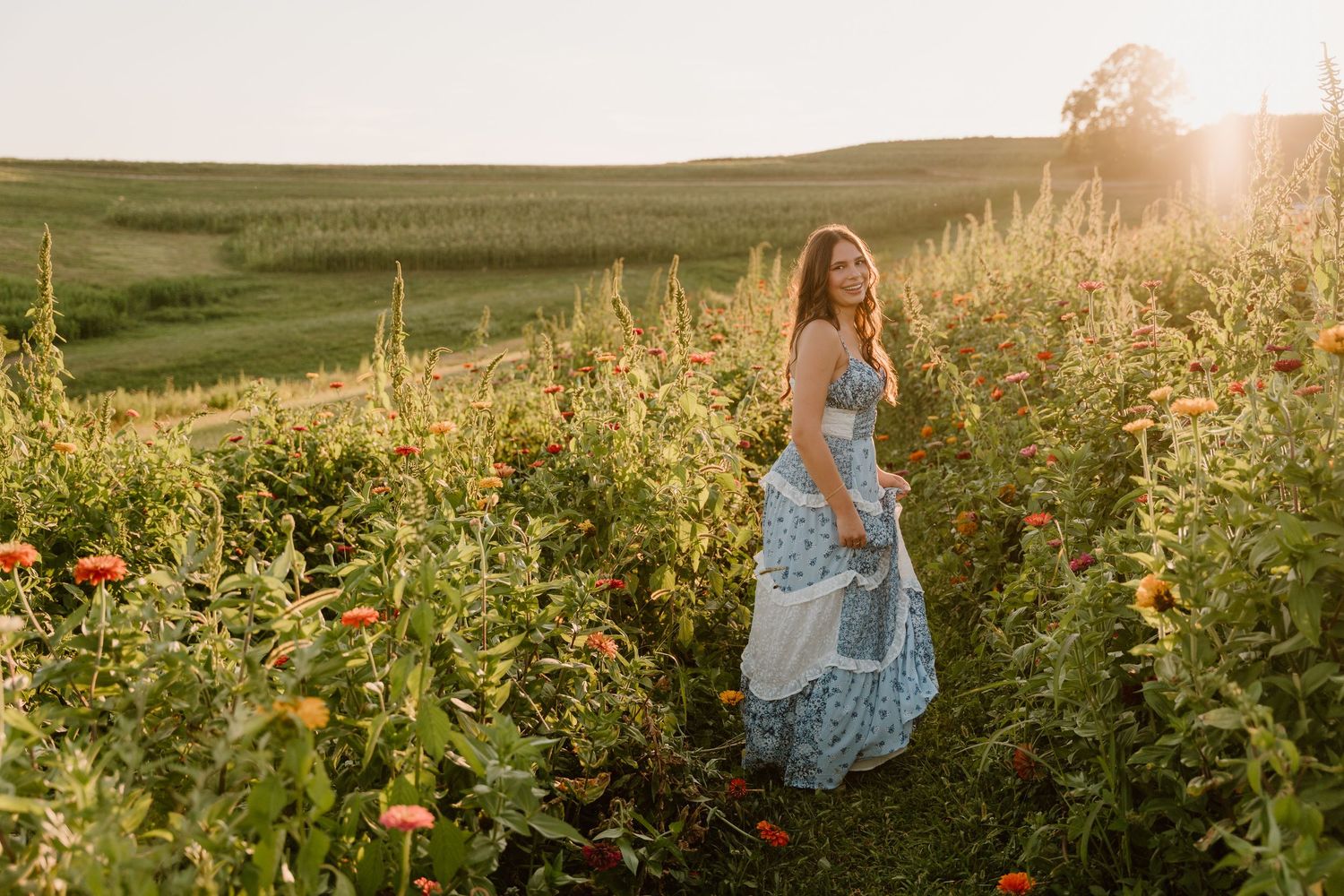 A blue floral dress flows in a golden sunlit field of wildflowers and tall grass during sunset.