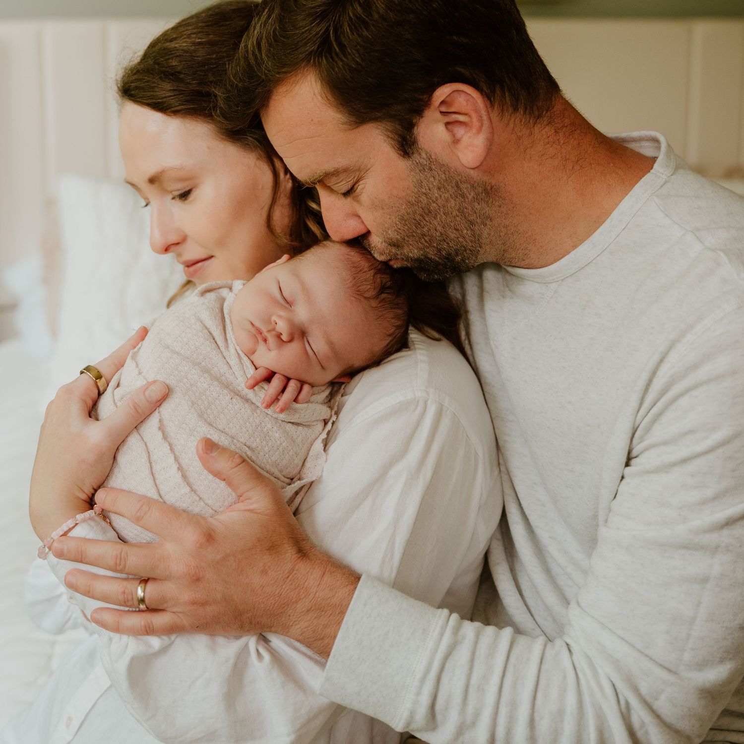 Tender family moment as parents in white clothing embrace their newborn baby in a bright, airy room.
