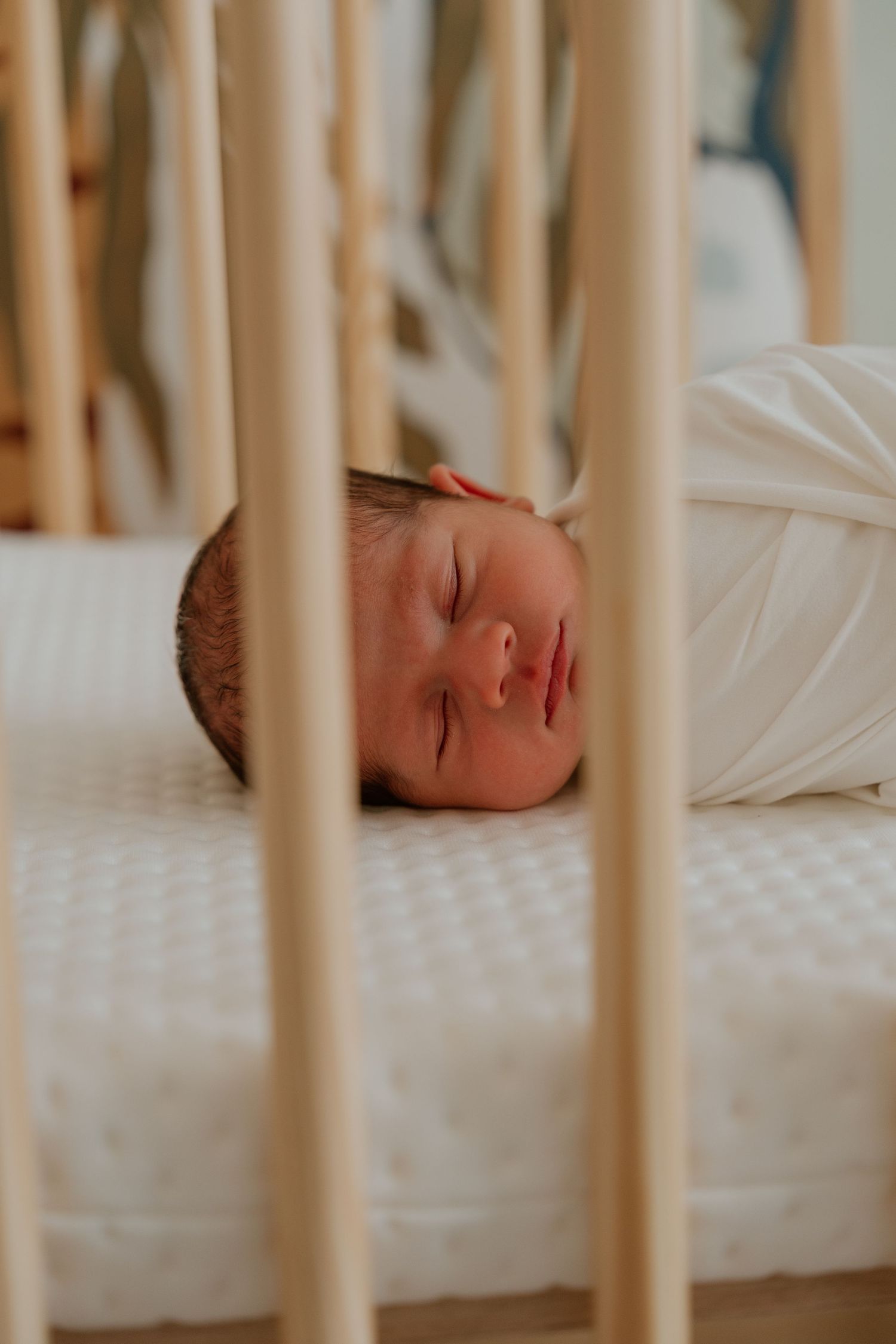 A sleeping baby lies peacefully in a white crib viewed through wooden slats.