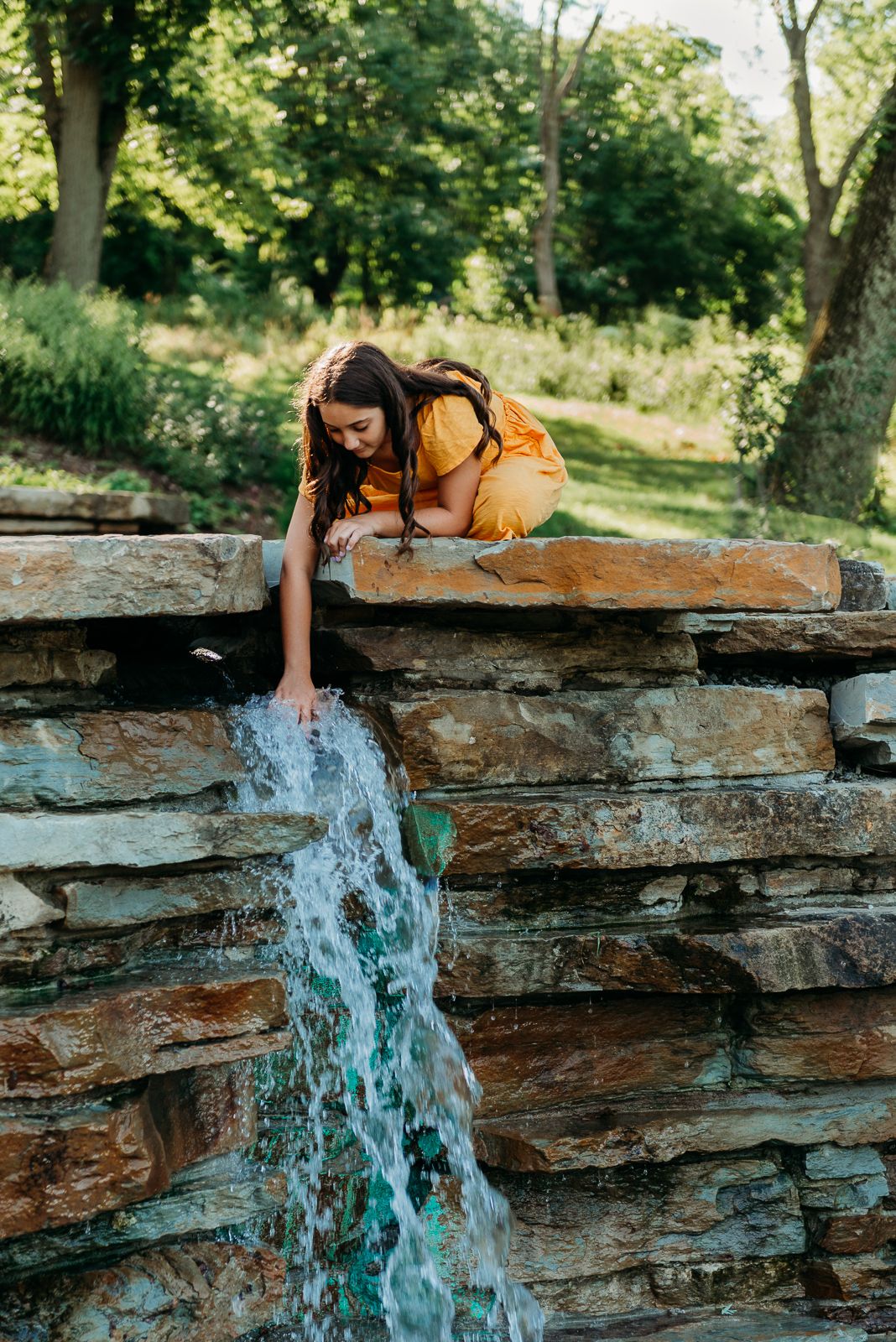 Person in yellow shirt reaching to touch waterfall cascading over layered rock formation.