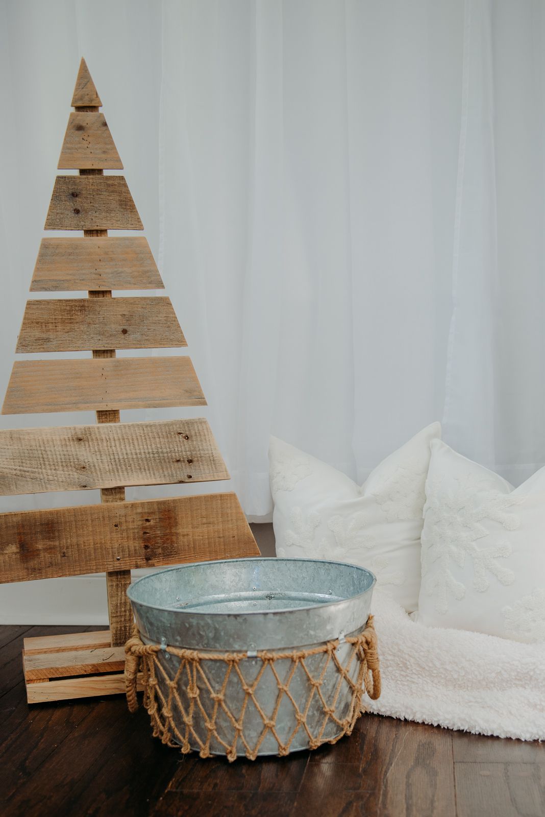 Rustic wooden triangle Christmas tree decoration with blue metal basket and white pillows on hardwood floor.