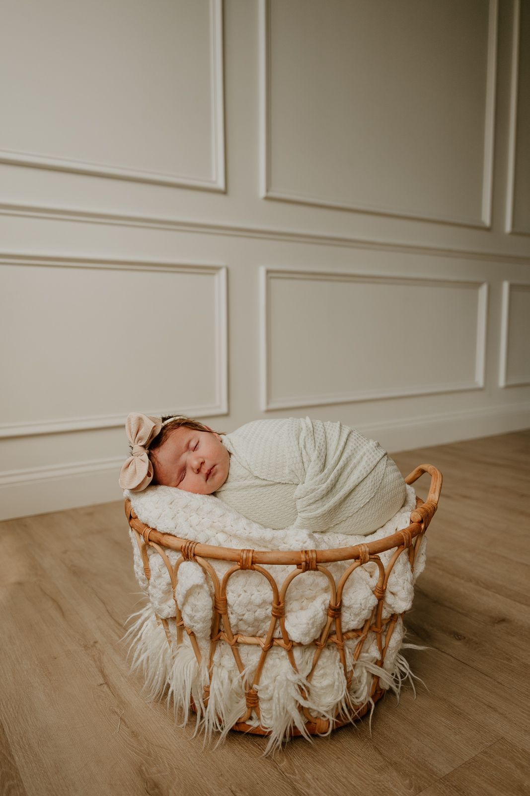 A sleeping newborn swaddled in white lies peacefully in a round bamboo basket with fluffy white trim on hardwood flooring.
