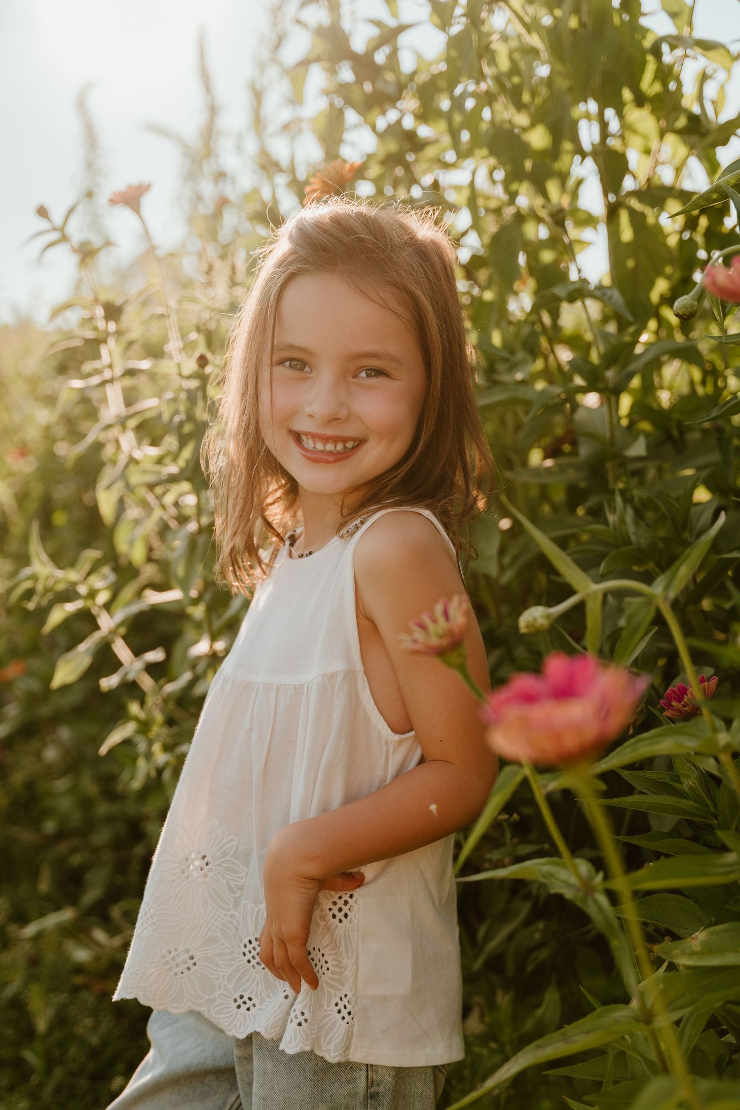 A smiling young girl in a white eyelet top stands among lush green plants and pink peonies in golden sunlight.