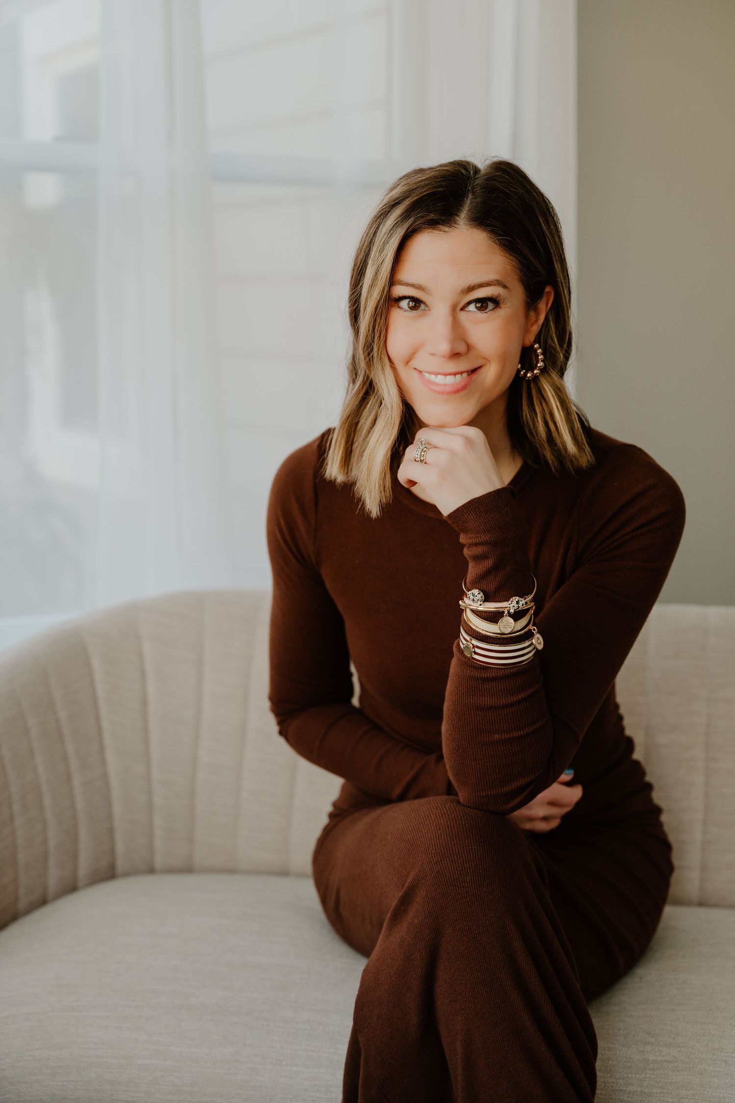 Woman in brown sweater dress sits comfortably on light gray couch in natural window light.